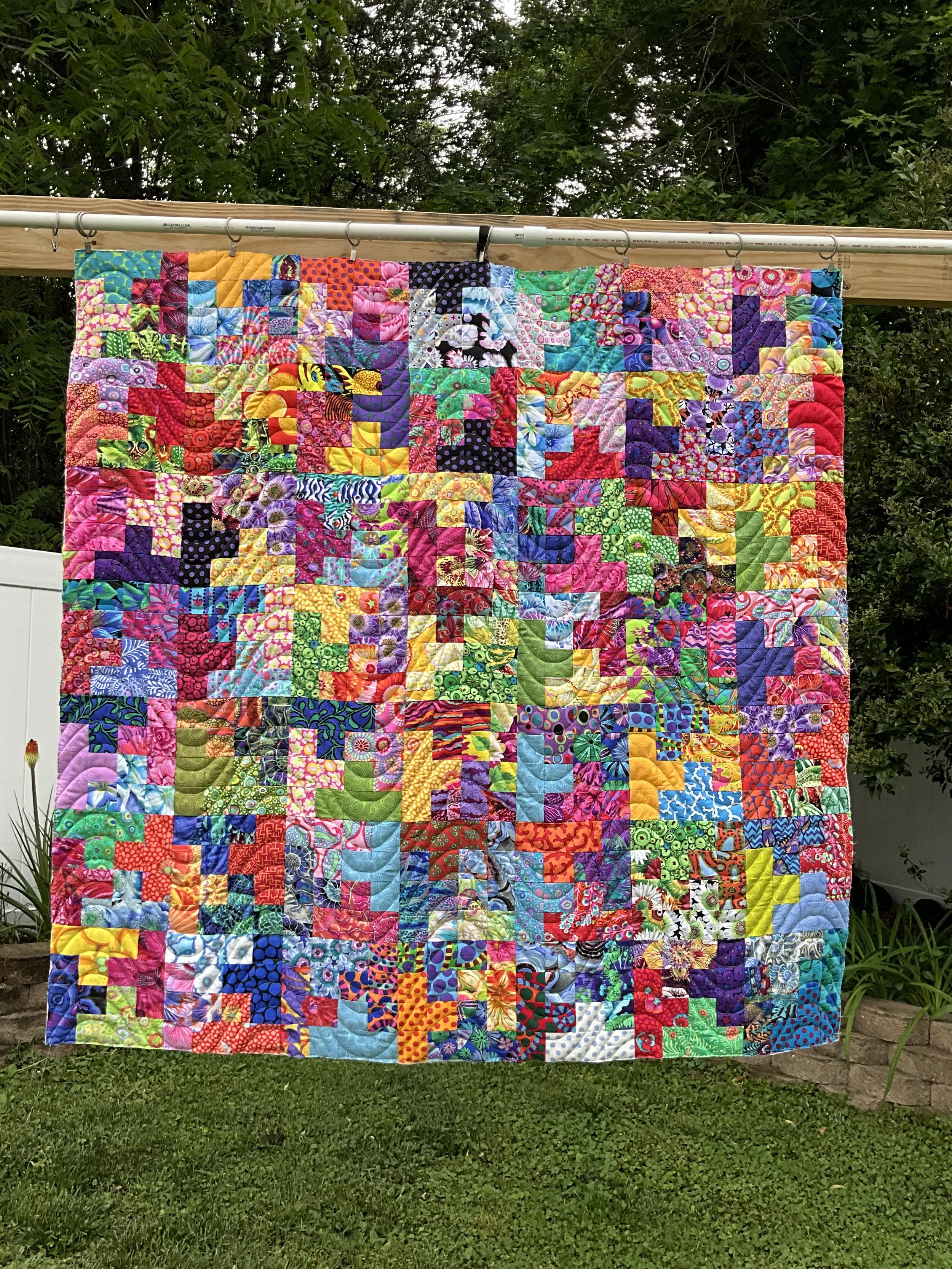 Colorful patchwork quilt hanging from a wooden clothesline outdoors with trees and greenery in the background.