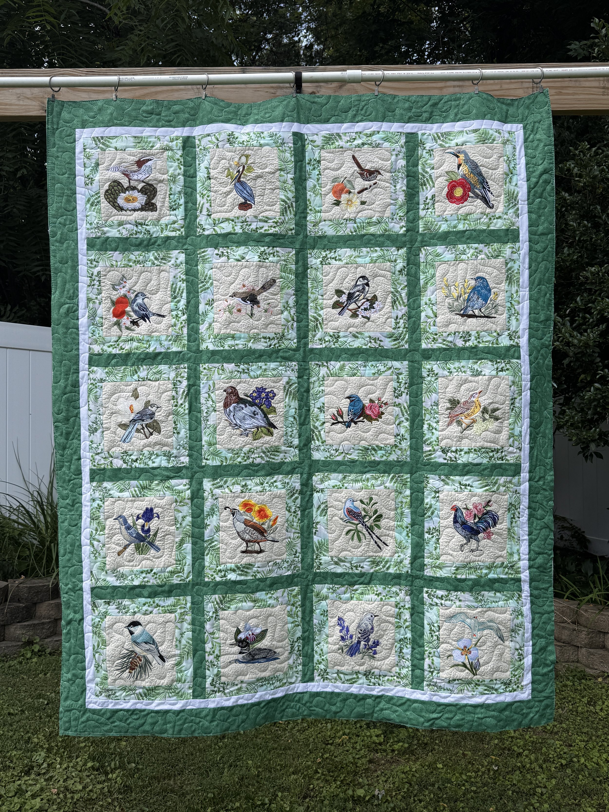 A quilt with embroidered bird designs, featuring various types of birds, bordered by green fabric, hanging outdoors on a wooden rod.