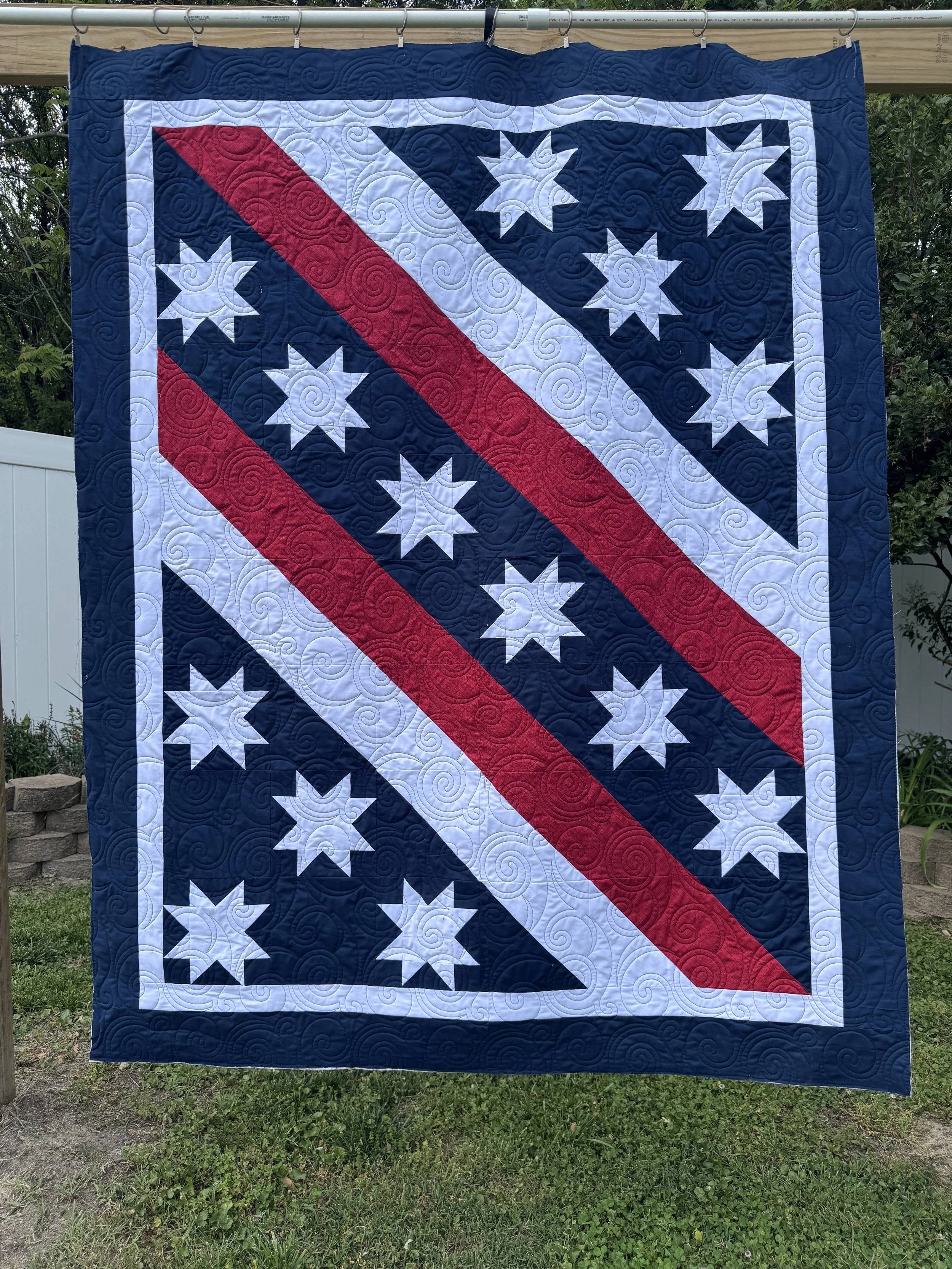 A quilt hanging outdoors on a clothesline, featuring a patriotic design with white stars on a blue background, red and white diagonal stripes, and a border of blue with swirl quilt patterns.