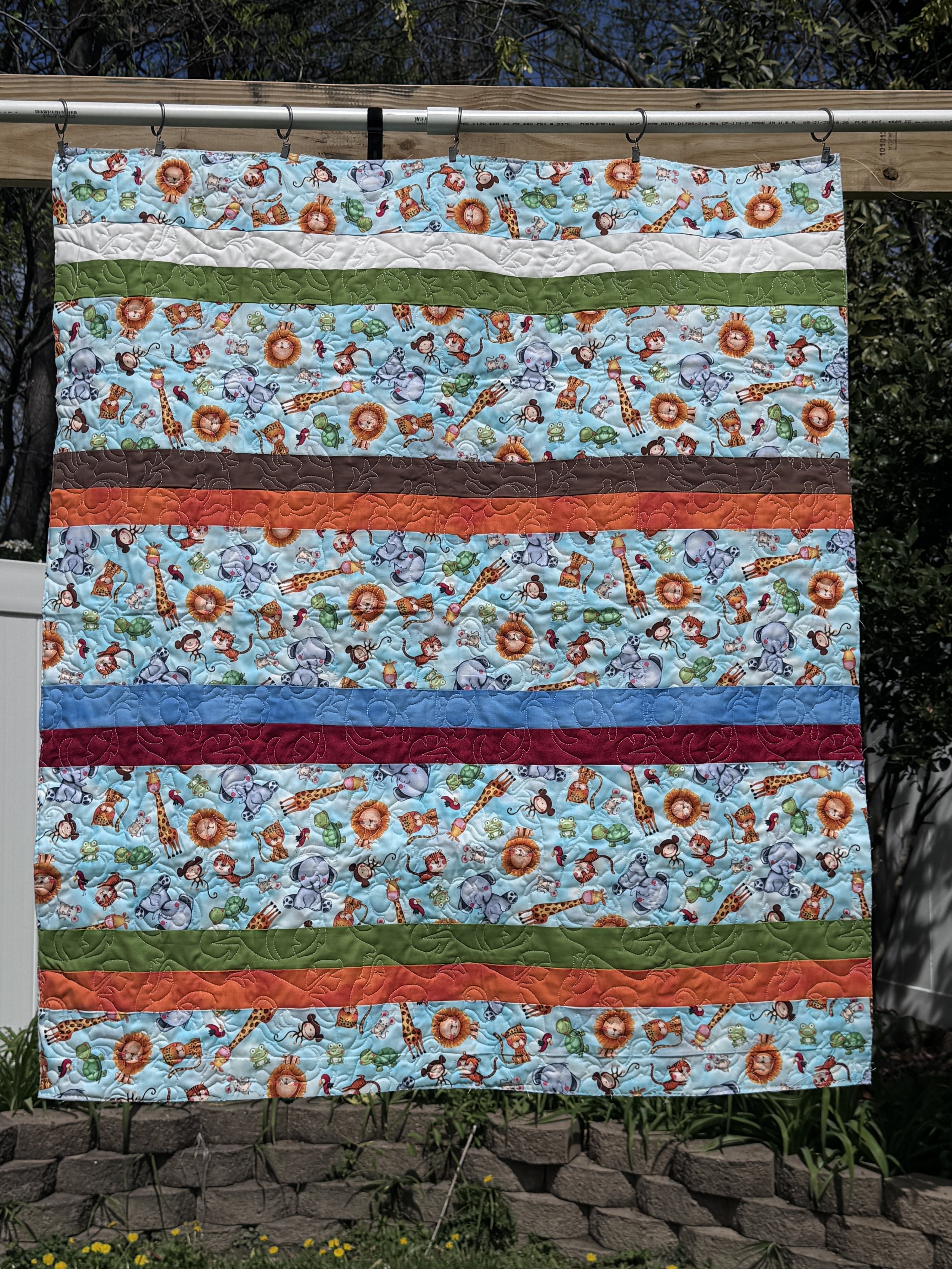 Colorful quilt with animal and jungle print, hanging outdoors on a clothesline with a brick garden border and plants in the background.