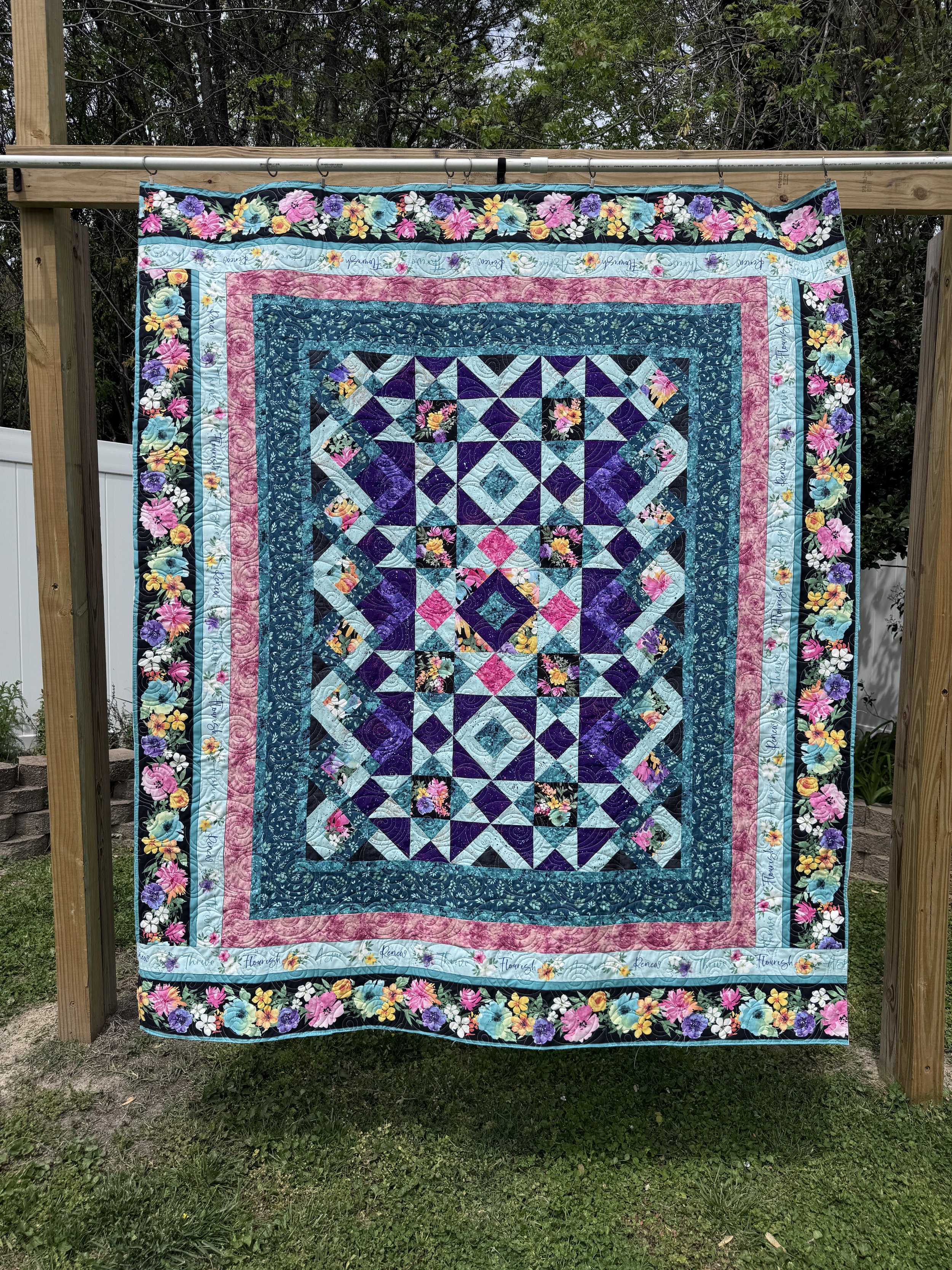 Colorful quilt hanging outdoors on a wooden frame, with a floral and geometric pattern in shades of pink, purple, blue, black, and white.