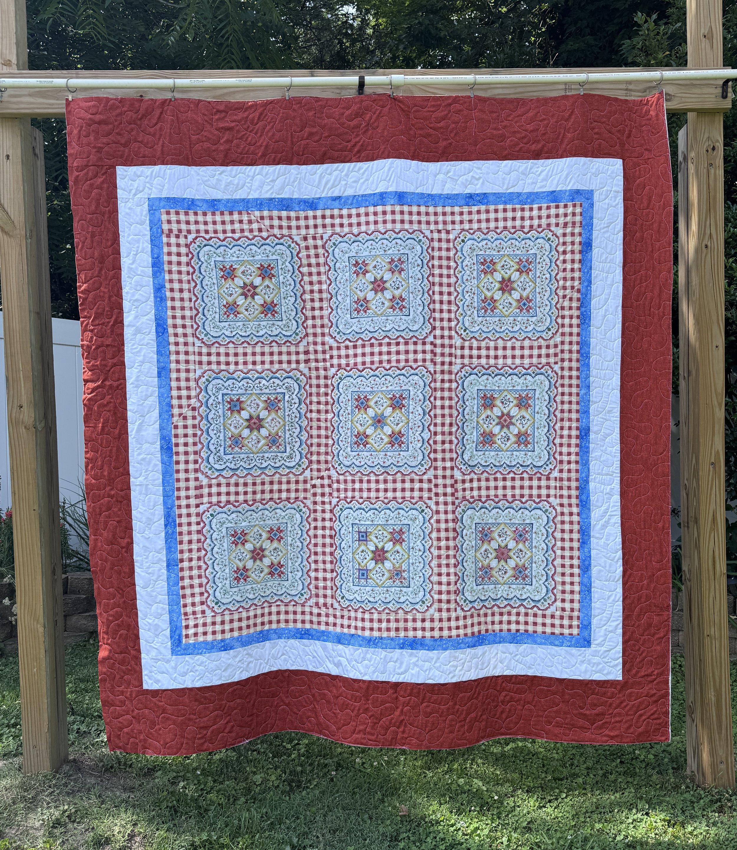 Colorful quilt hanging outdoors on a wooden frame with trees in the background. The quilt has a red outer border, a white inner border, and a central pattern of embroidered squares with intricate designs and red checkered backgrounds.