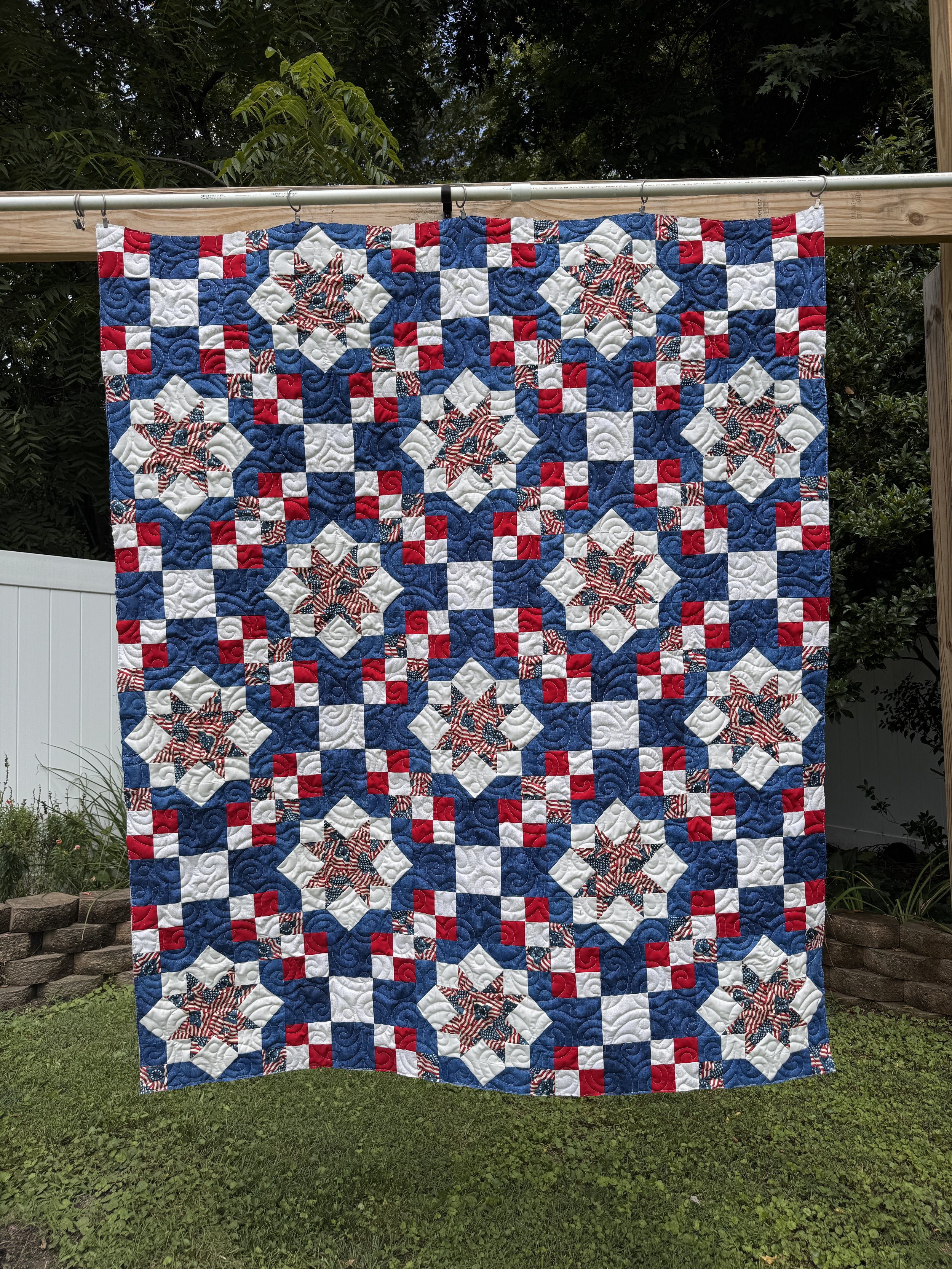 A patriotic quilt hanging outdoors on a wooden frame with trees in the background.