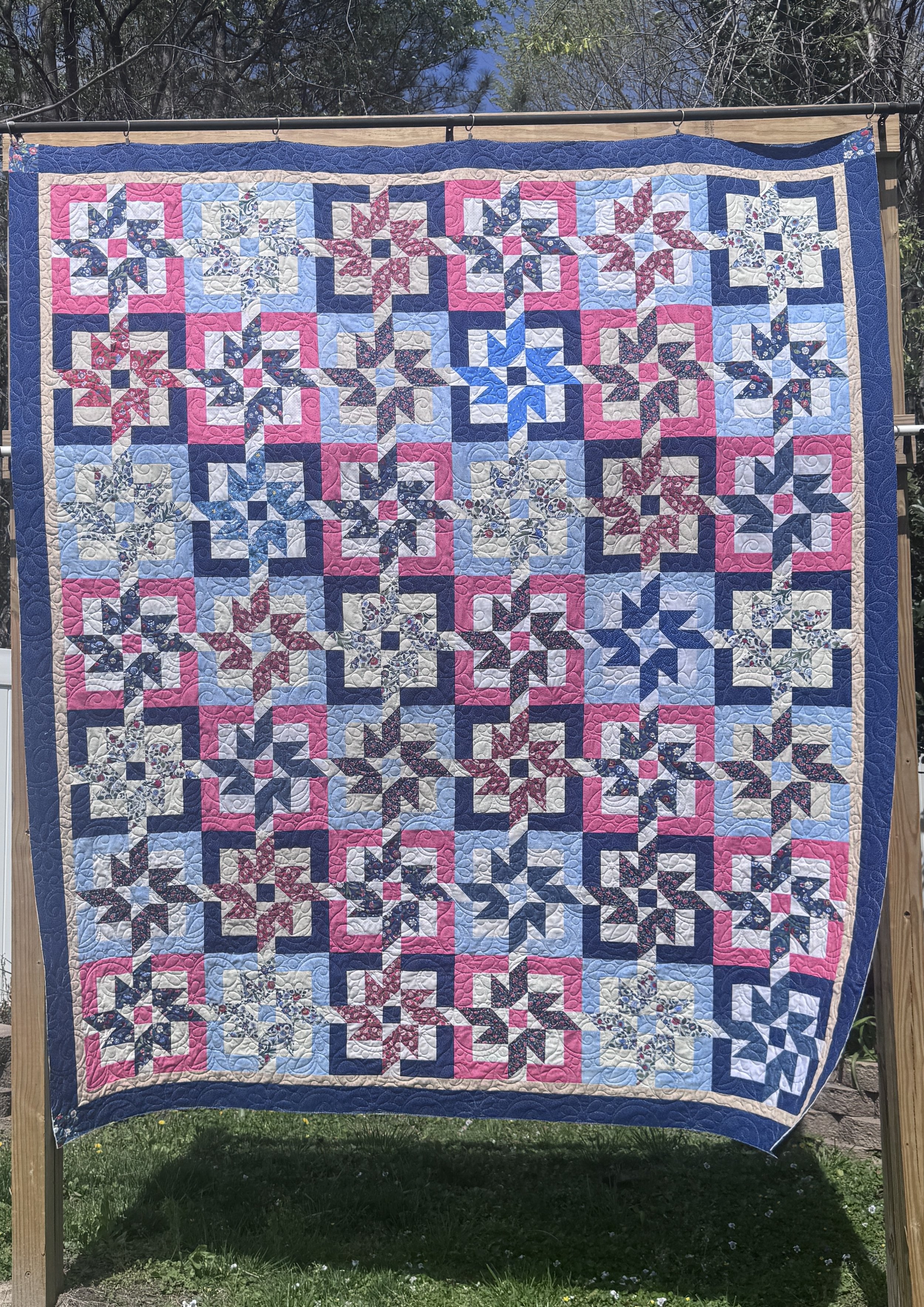 A colorful quilt hanging on a wooden frame outdoors, featuring star patterns in shades of pink, blue, white, and floral designs with a blue border.
