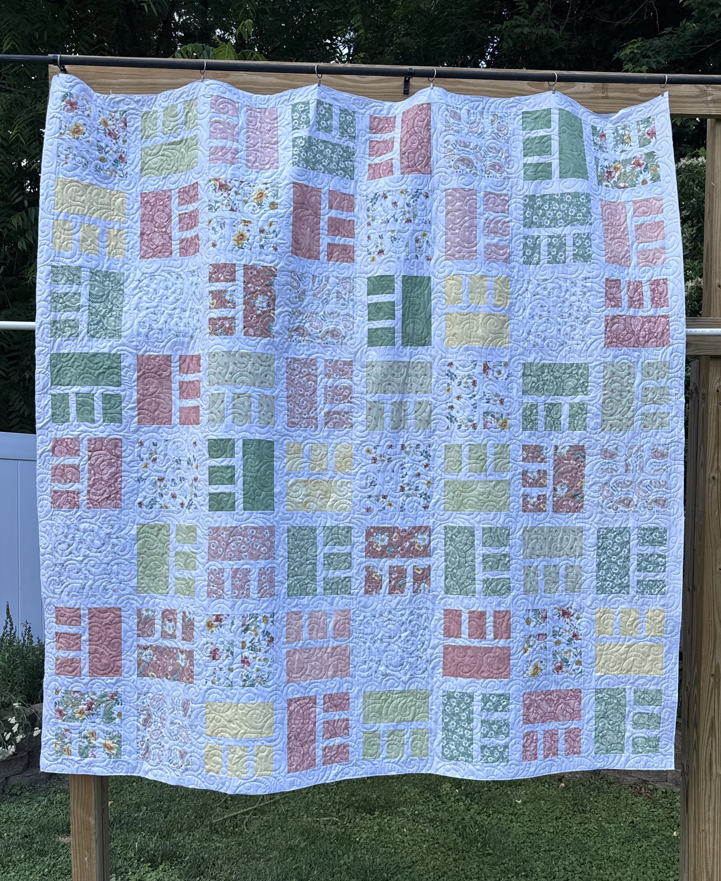 A quilt hanging outdoors on a wooden frame, with pastel-colored geometric and floral patterns, surrounded by green trees and grass.
