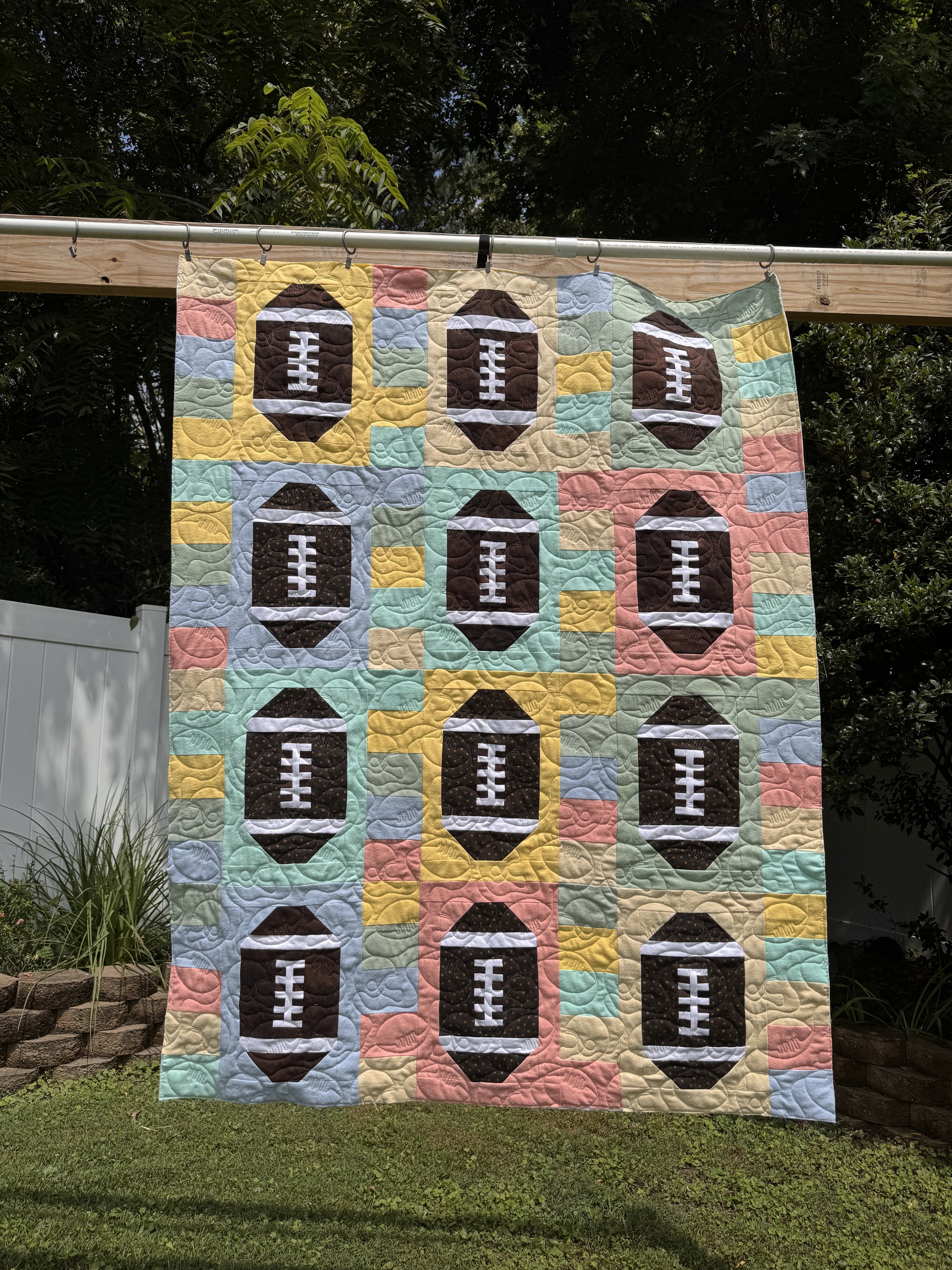 A colorful quilt featuring football patterns hangs on a line outdoors, with green trees and a white fence in the background.