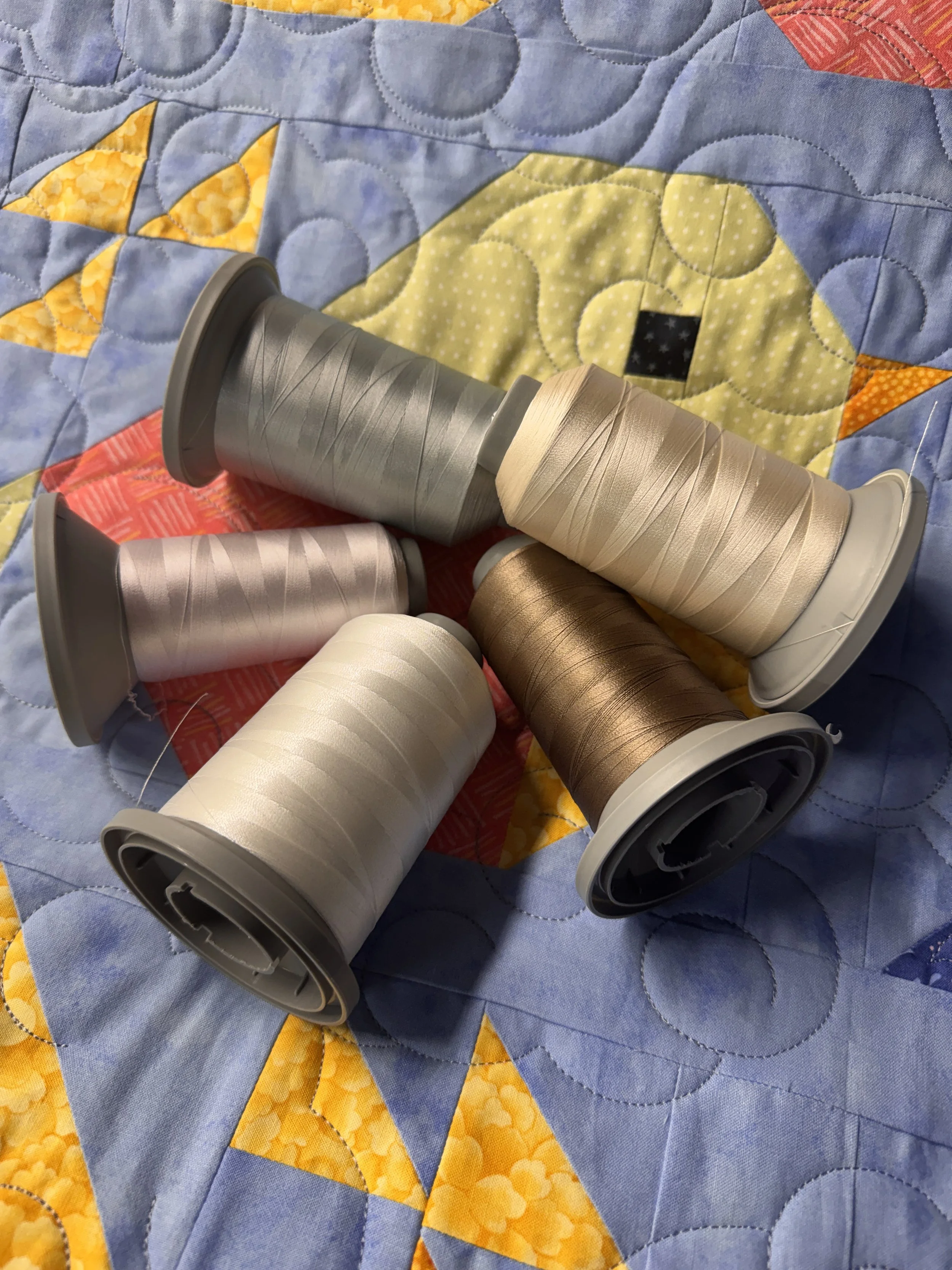 Several spools of metallic-colored sewing thread in shades of silver, gold, bronze, white, and pink are placed on a colorful quilt with a flower pattern, including yellow, orange, and blue sections.