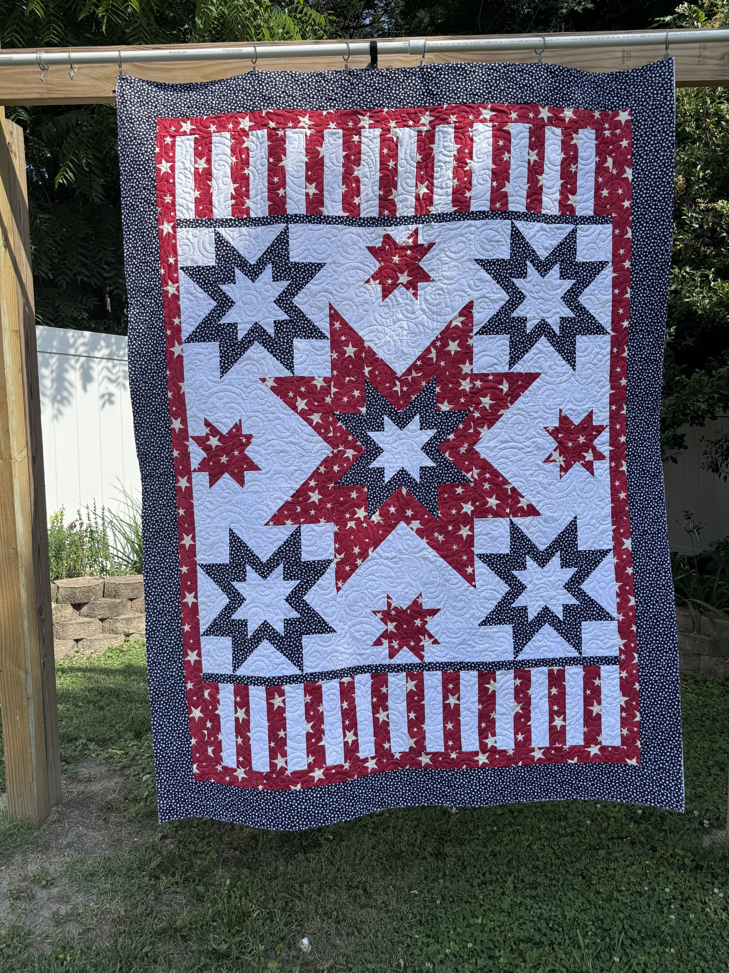 A patriotic quilt hanging on a horizontal pole outdoors. The quilt features red, white, and blue colors with star and stripe patterns reminiscent of the American flag.