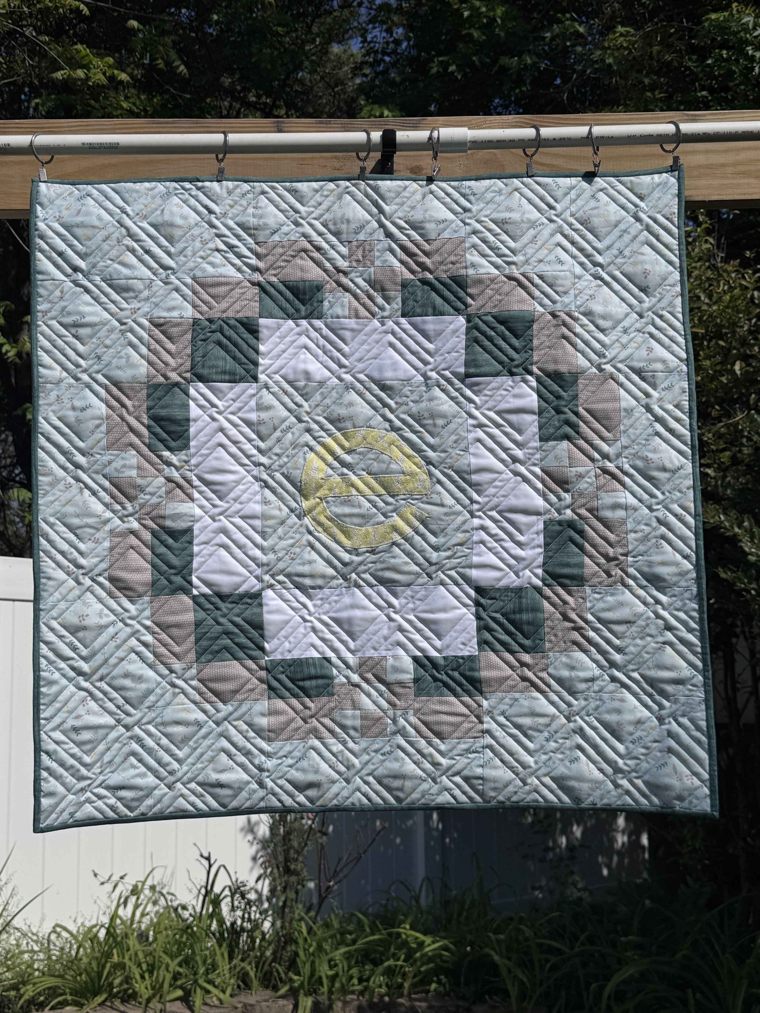 A quilt hanging outside on a metal rod, with a nature scene background of trees and partially cloudy sky.
