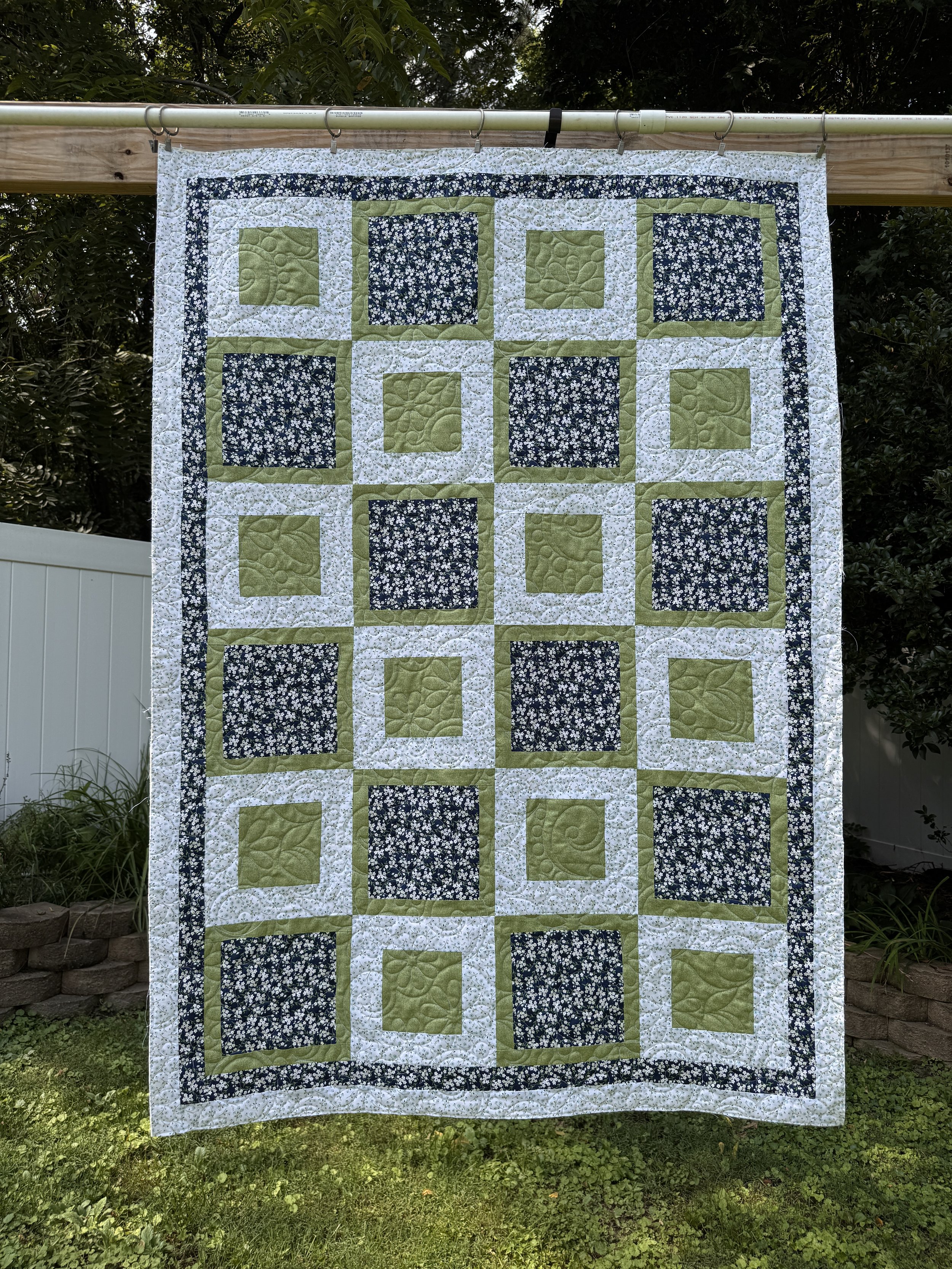 A quilt with a patchwork design of alternating green and blue floral patterns, bordered by a white floral fabric, hanging outdoors on a clothesline in a garden.