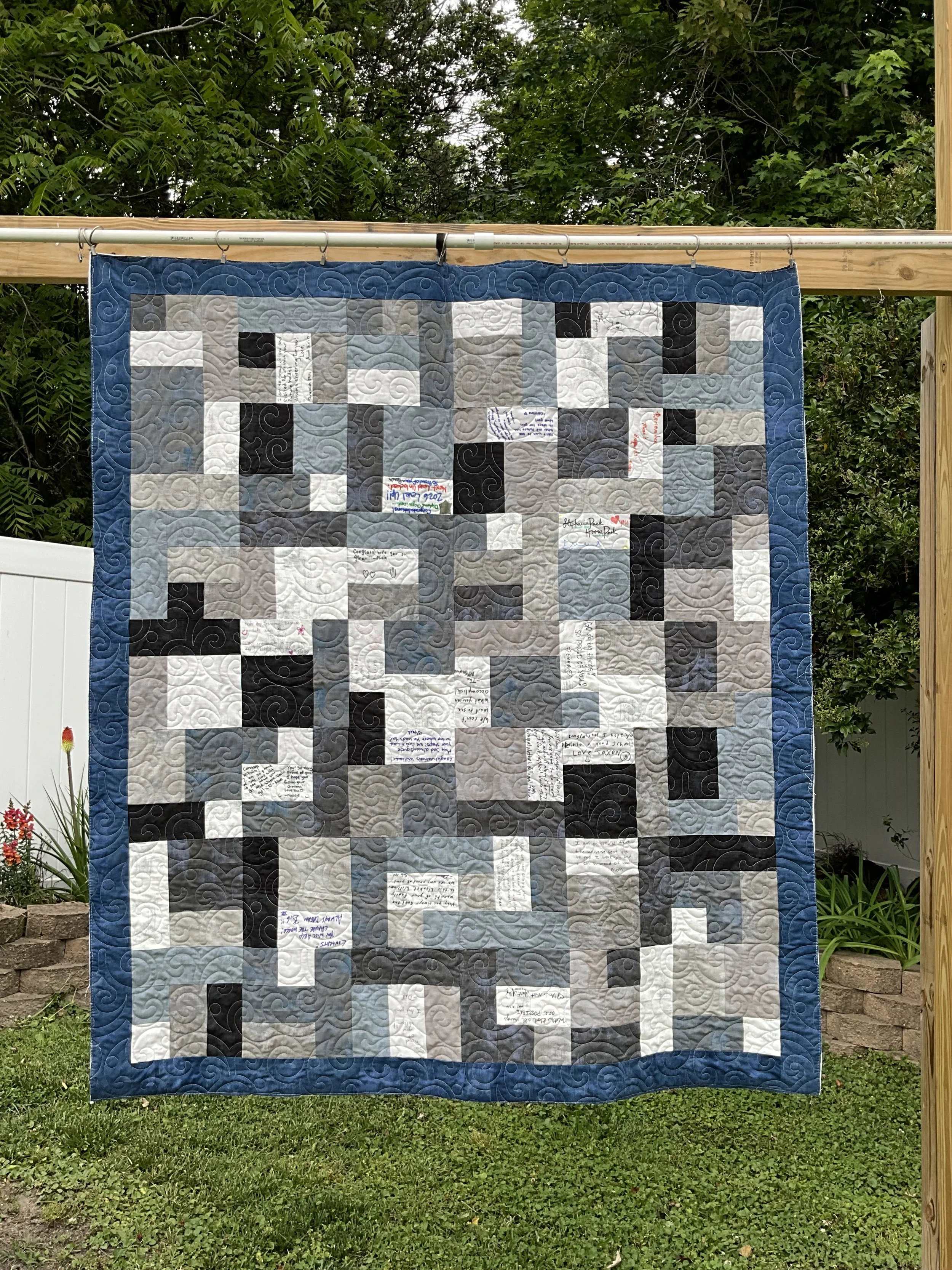 A quilt hanging on a clothesline in a garden with green trees and plants in the background. The quilt has a patchwork pattern with shades of black, white, gray, and blue, and contains handwritten notes on some of the patches.