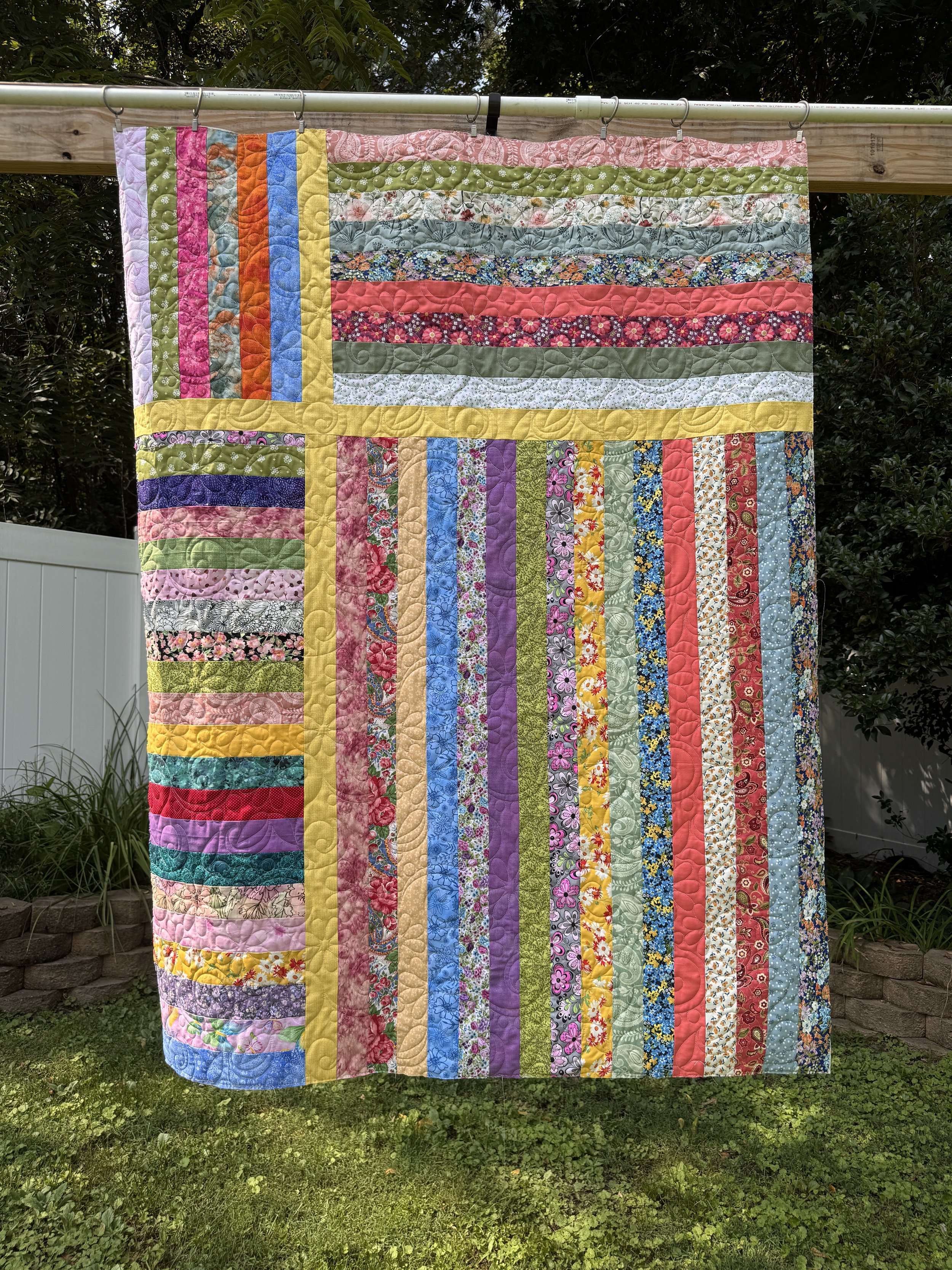 A colorful patchwork quilt hanging outdoors on a clothesline, surrounded by green plants and trees.