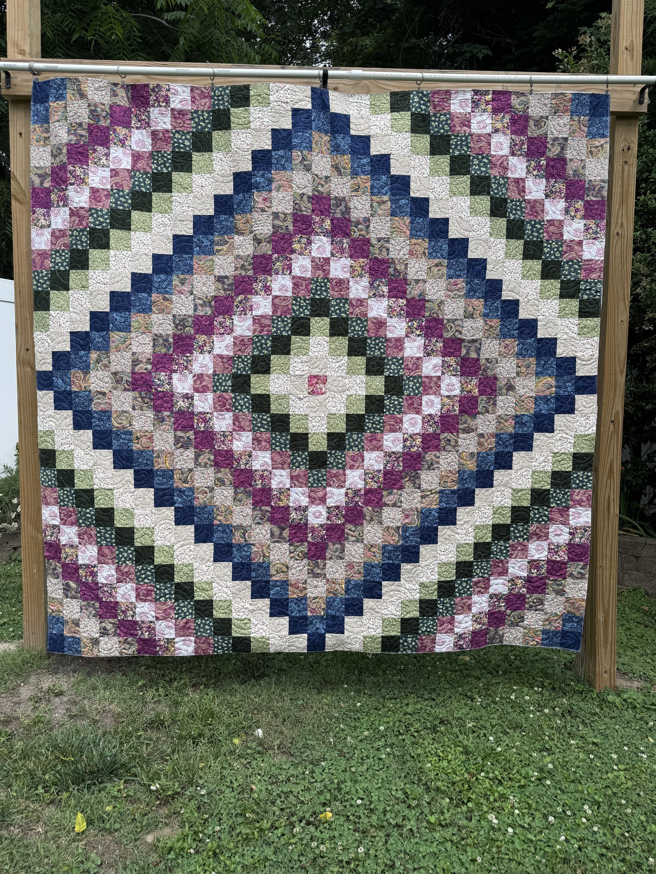 A colorful quilt featuring a symmetrical floral pattern in shades of purple, blue, green, white, and beige, hanging on a wooden frame outdoors in a grassy area with trees in the background.