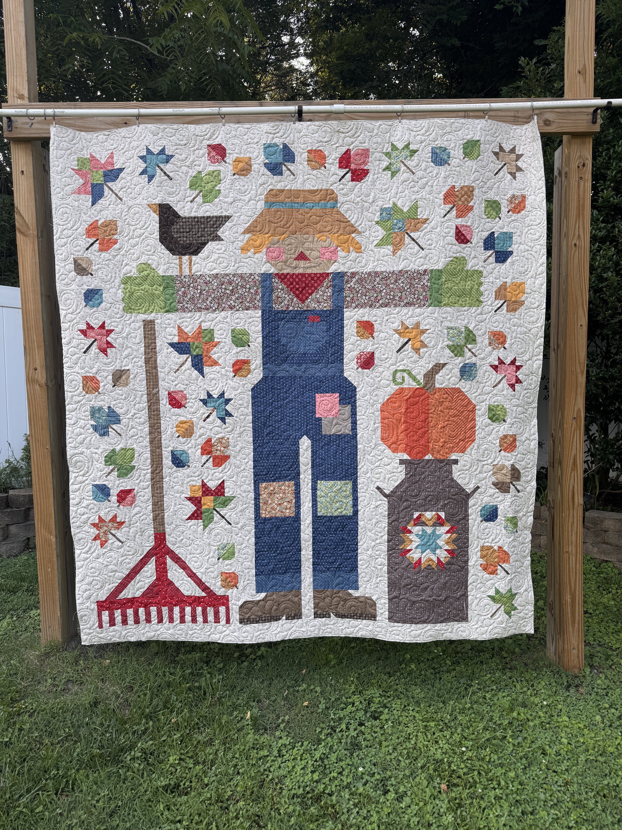A quilt hanging on a wooden frame, featuring a scarecrow surrounded by autumn leaves and a pumpkin. The scarecrow wears a straw hat, a red bandana, and blue overalls. The quilt has intricate quilting patterns and colorful fabric pieces against a whit