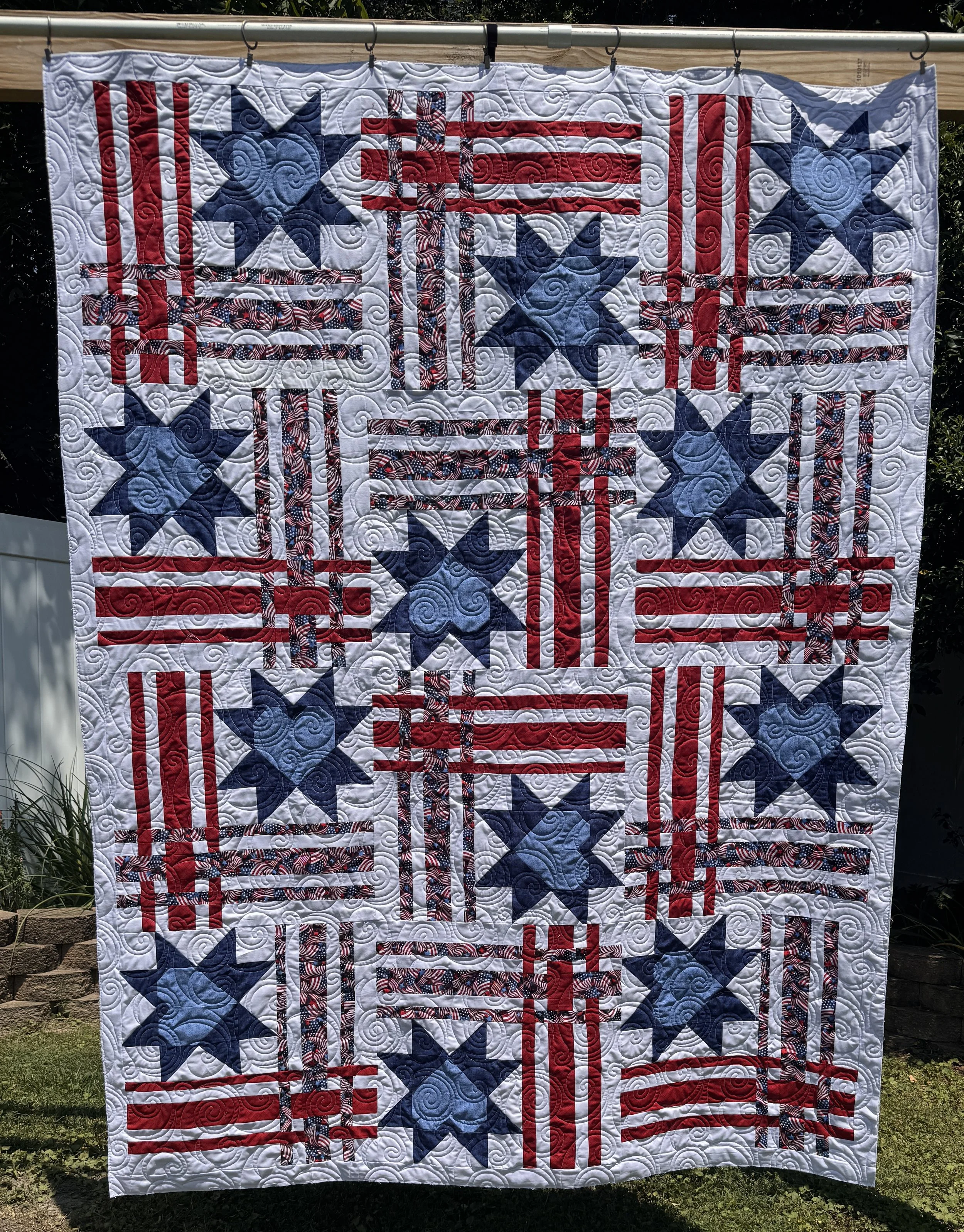 A quilt with patriotic American themes, featuring stars and stripes in red, white, and blue patterns, hanging outdoors on a wooden rod.