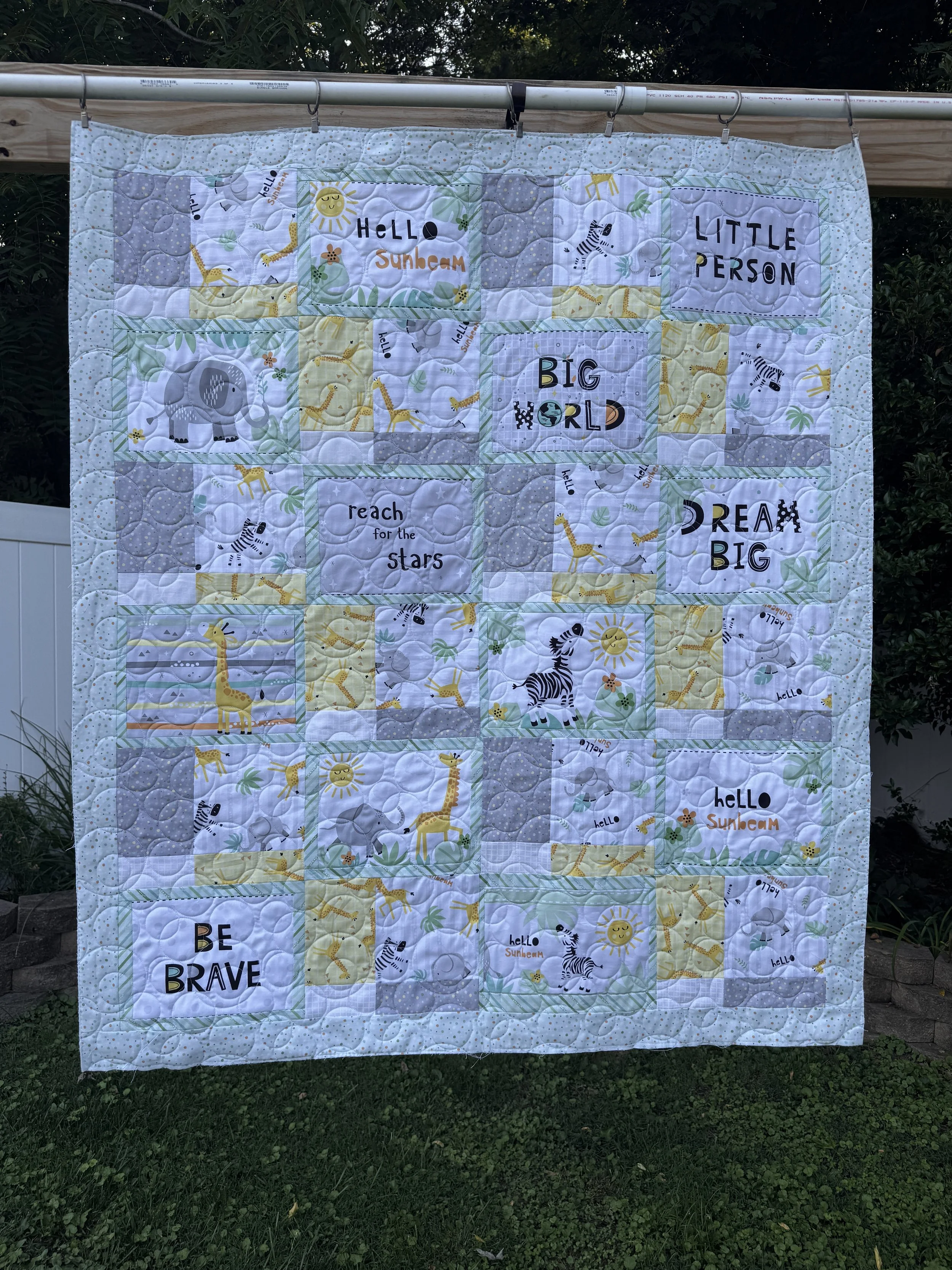 A quilt hanging outdoors on a horizontal rod with a background of trees and grass. The quilt features blocks with animal illustrations like a giraffe, zebra, and elephant, and contains words such as 'HELLO,' 'SUNSHINE,' 'LITTLE PERSON,' 'BIG WORLD,' 