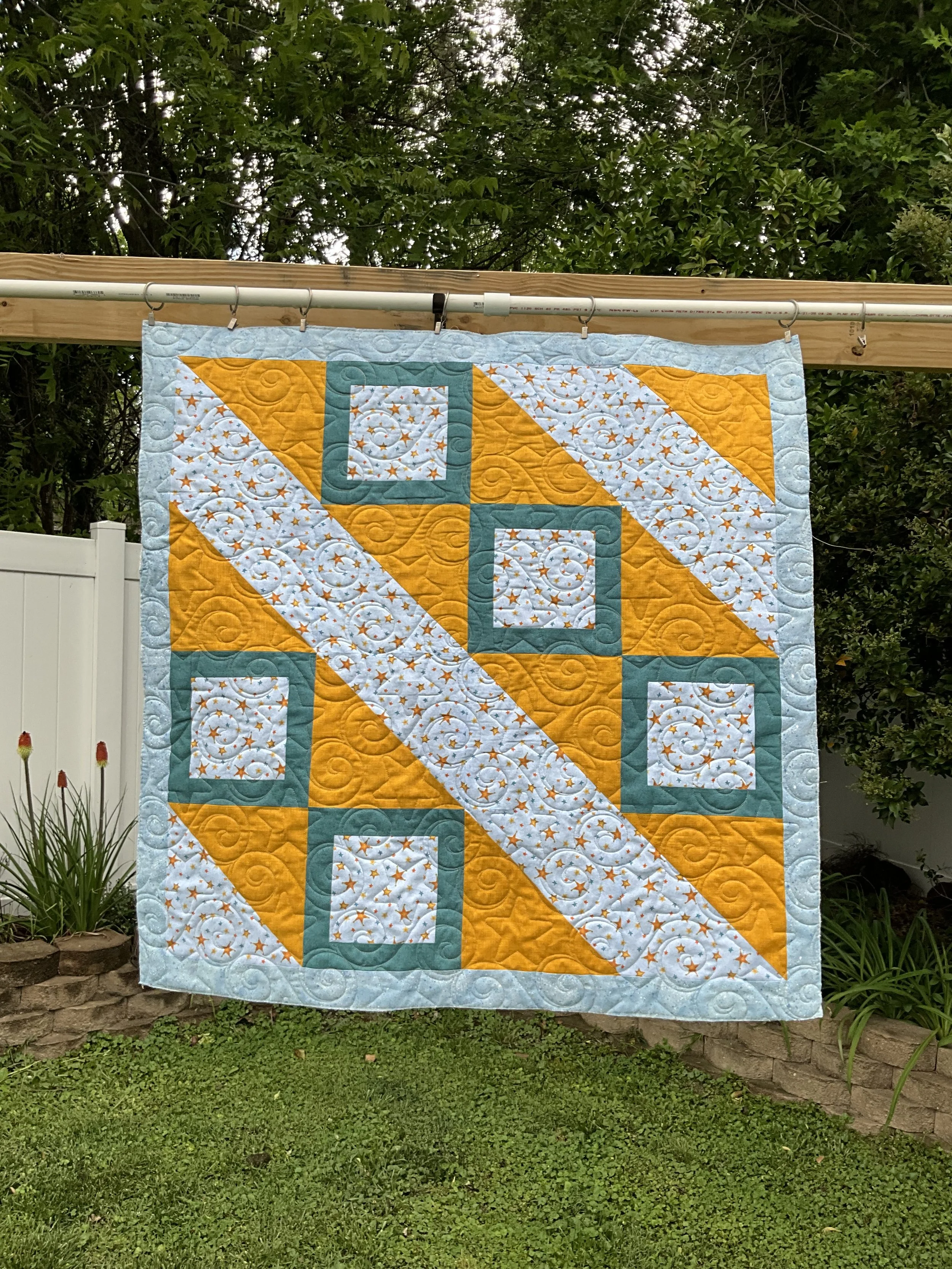 Colorful quilt hanging outdoors on a wooden pole, with trees and bushes in the background and green grass below.