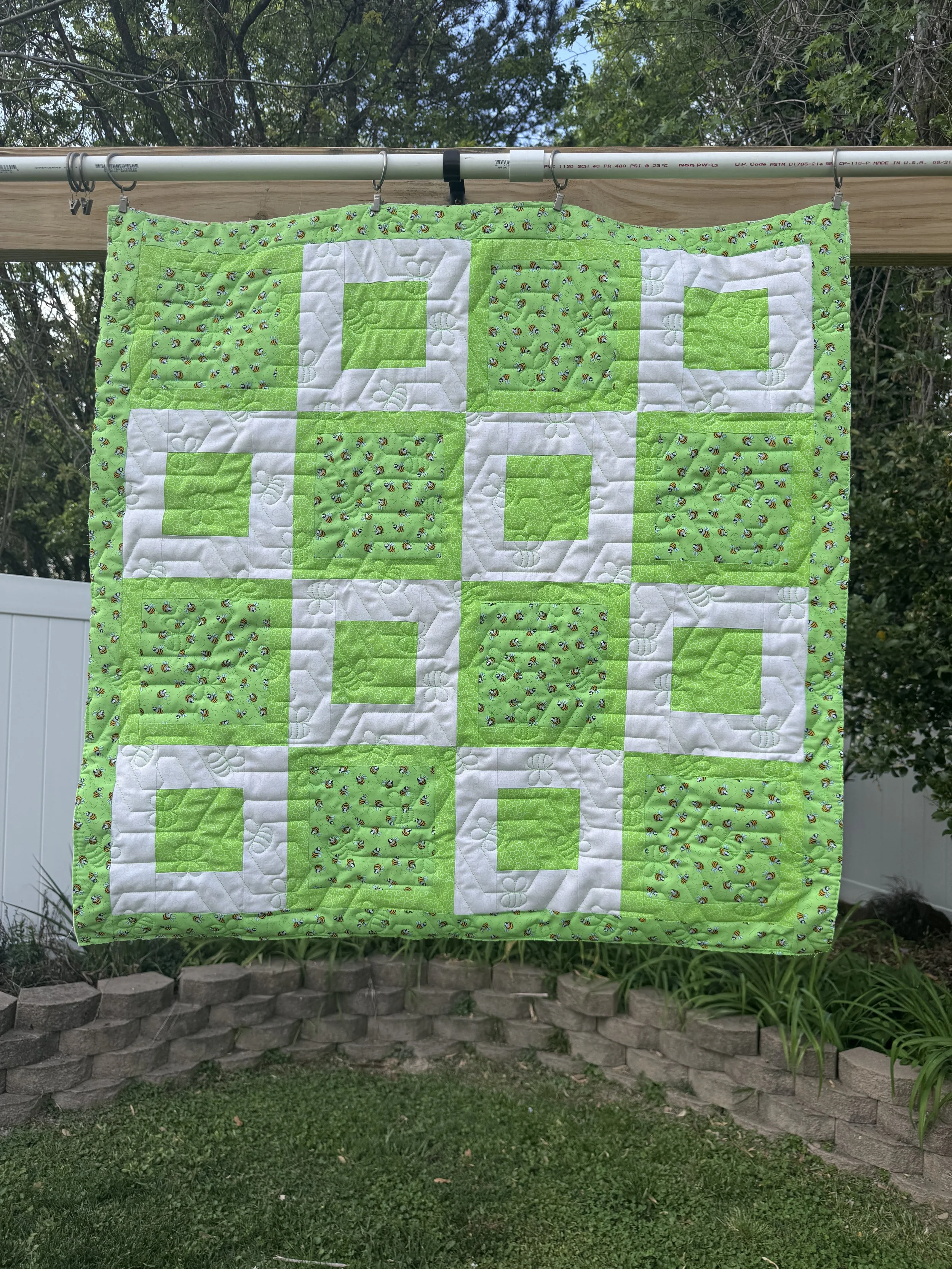 Green and white quilt with bee and flower patterns hanging outdoors on a clothesline with trees and a brick garden bed in the background.