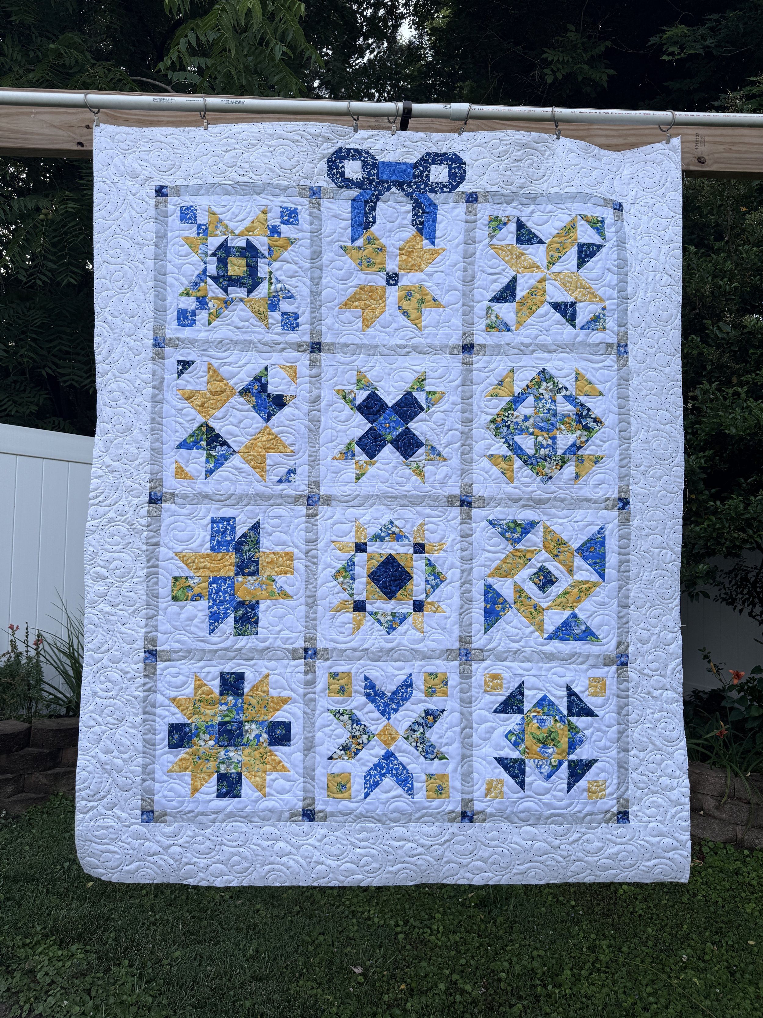 Handmade quilt with various patchwork blocks featuring blue, yellow, and white fabrics, hanging outdoors on a rod with greenery in the background.