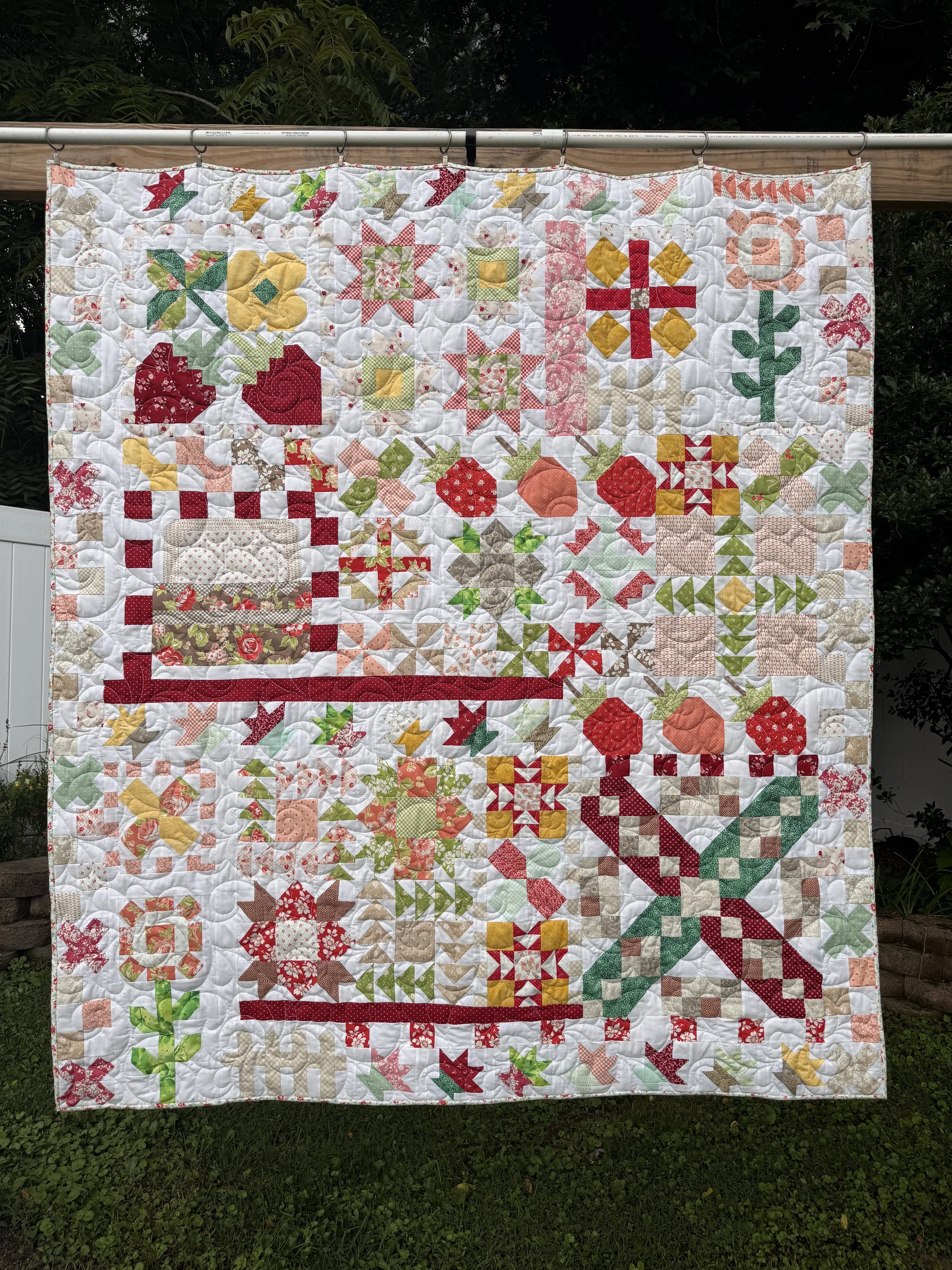 A colorful quilt hanging outdoors on a clothesline, featuring various Christmas-themed patterns such as holly, poinsettias, crosses, and stars in red, green, yellow, and beige colors.