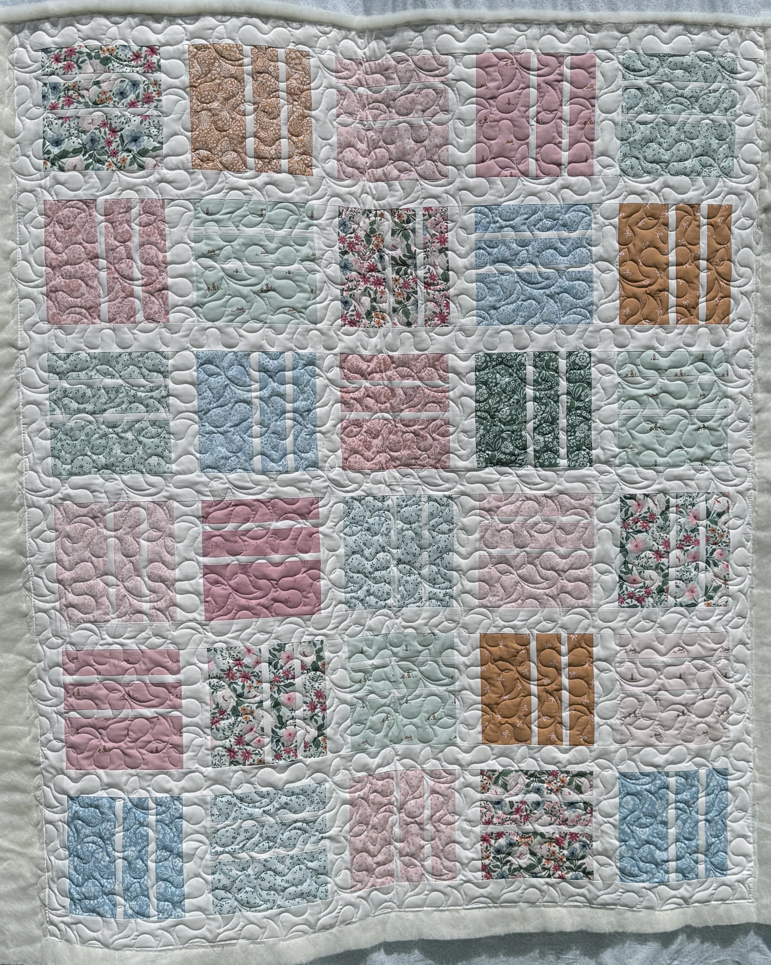 A quilt with a pattern of rectangular blocks, each containing different floral and geometric fabric designs in pastel pink, blue, green, brown, and white, with quilting stitches creating a wavy pattern across the surface.