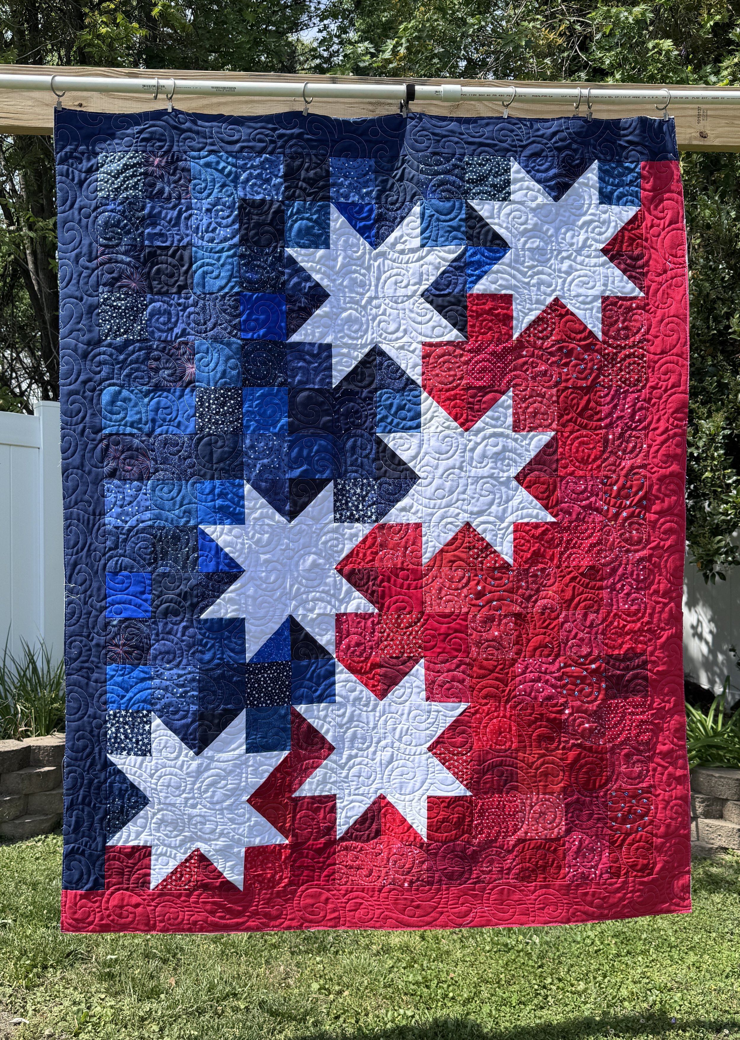 A quilt hanging outdoors with a patriotic color theme of red, white, and blue, featuring large star patterns and swirling quilted designs.