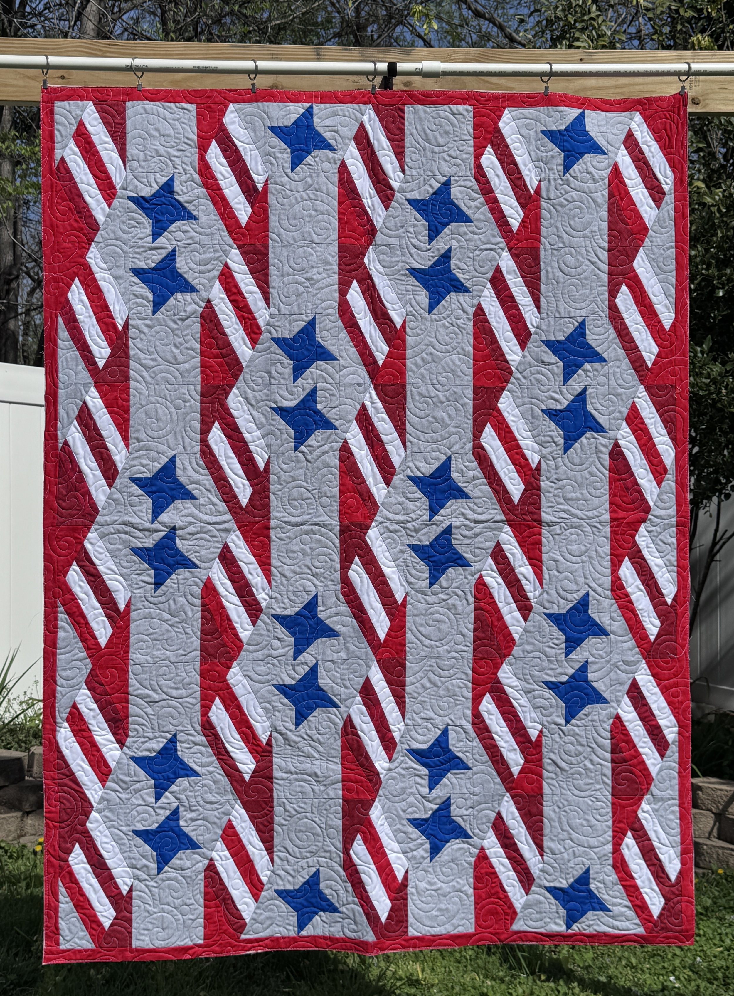 Square quilt quilted with red, white, gray, and blue colors. Features star patterns and diagonal stripes, hung outdoors against a wooden fence.