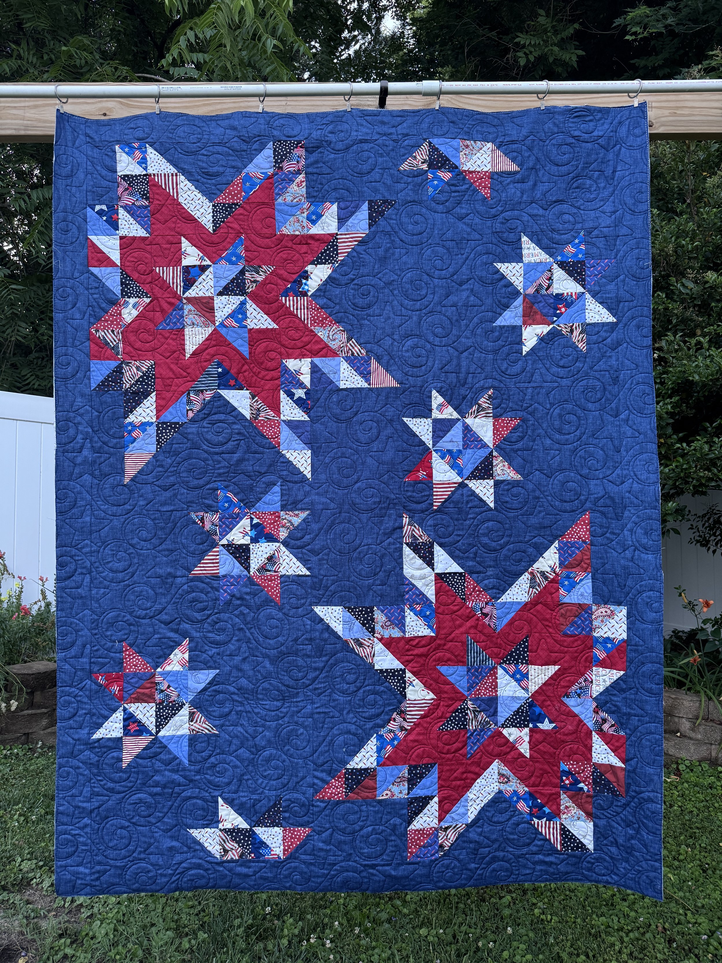 A patriotic quilt with stars and flag-patterned fabrics, featuring a large red star, multiple smaller stars, and quilted swirl patterns, hanging outdoors against green trees.