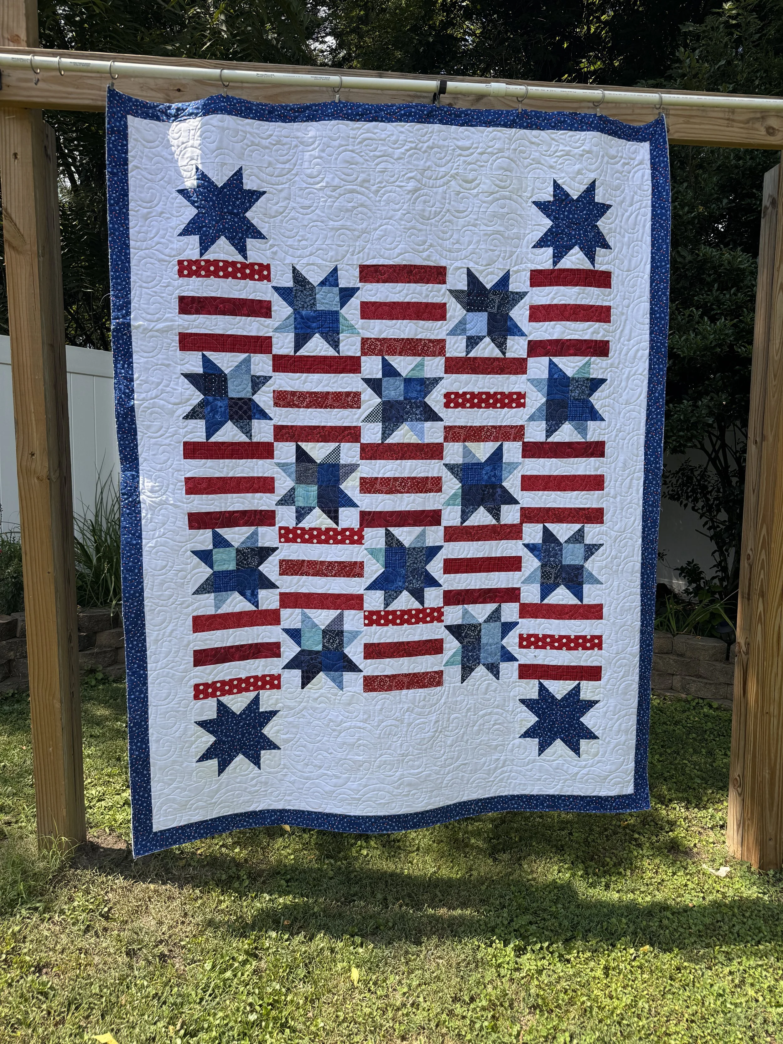 A quilt hanging on a wooden frame outdoors with a design inspired by the American flag, featuring red and white stripes and blue stars.