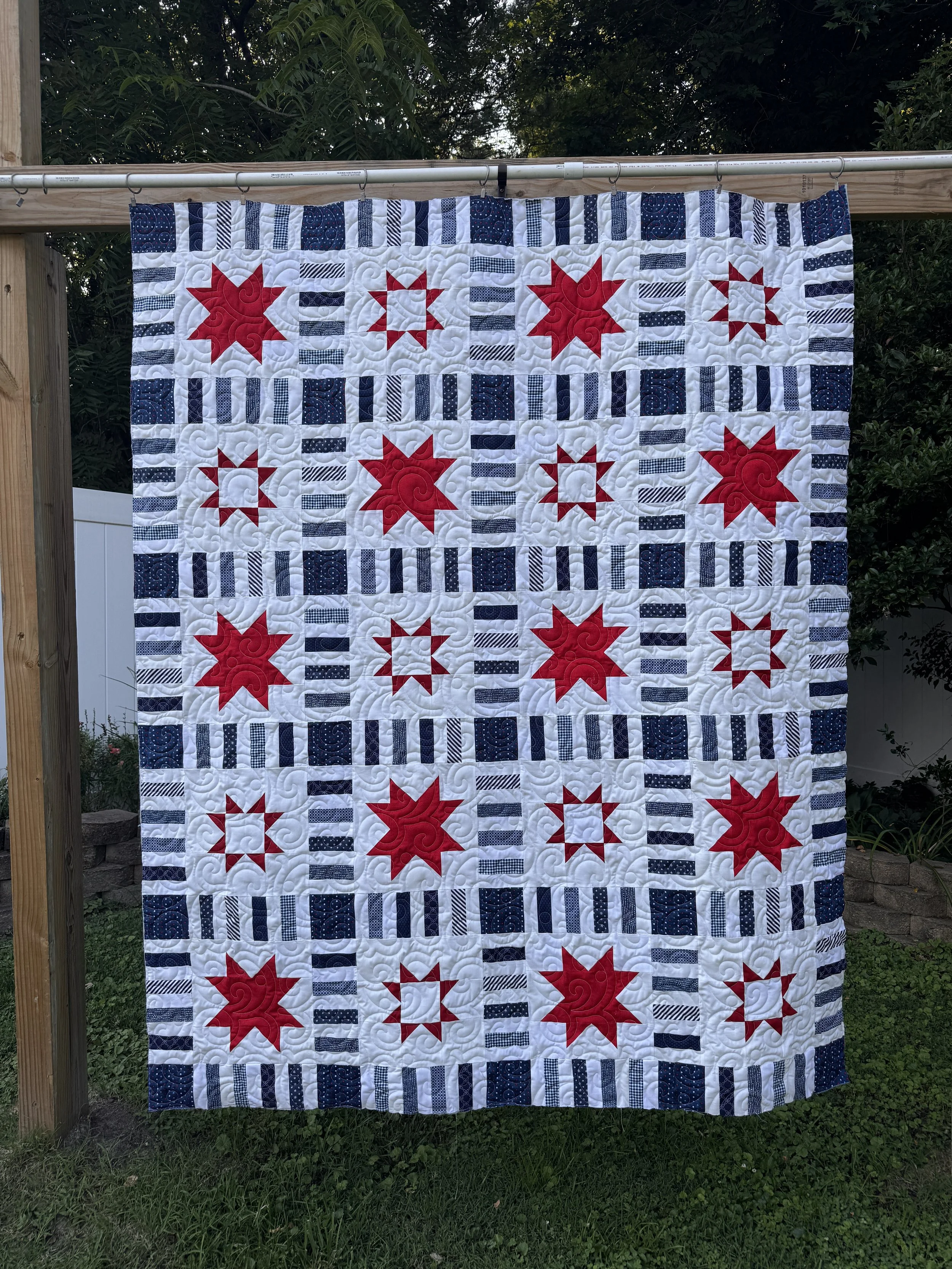 A quilt hanging outdoors on a clothesline between wooden posts. The quilt has a patriotic theme with red, white, and blue colors, featuring star patterns and striped designs.