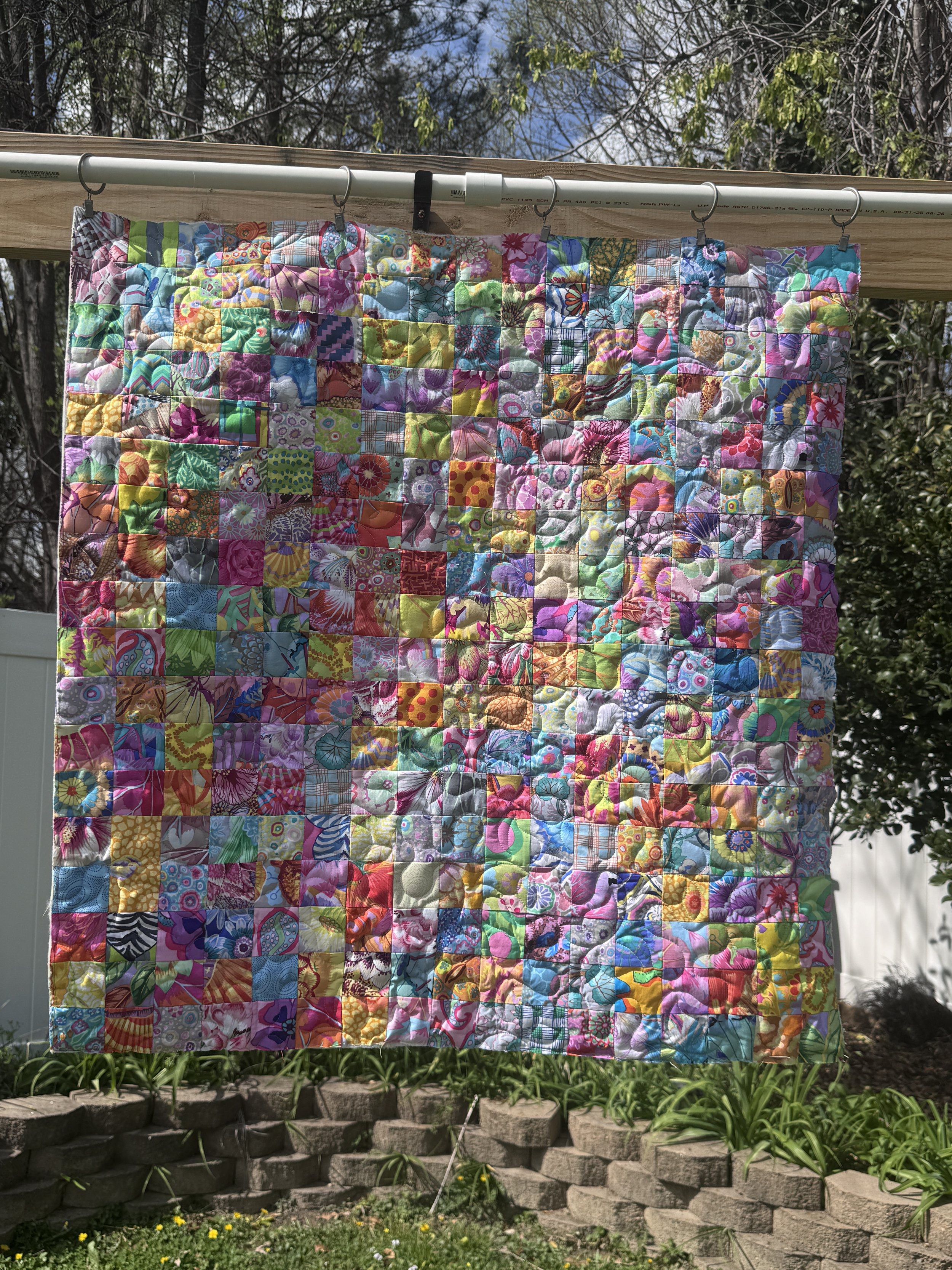 Colorful quilt hanging outdoors on a clothesline, with trees and blue sky in the background.