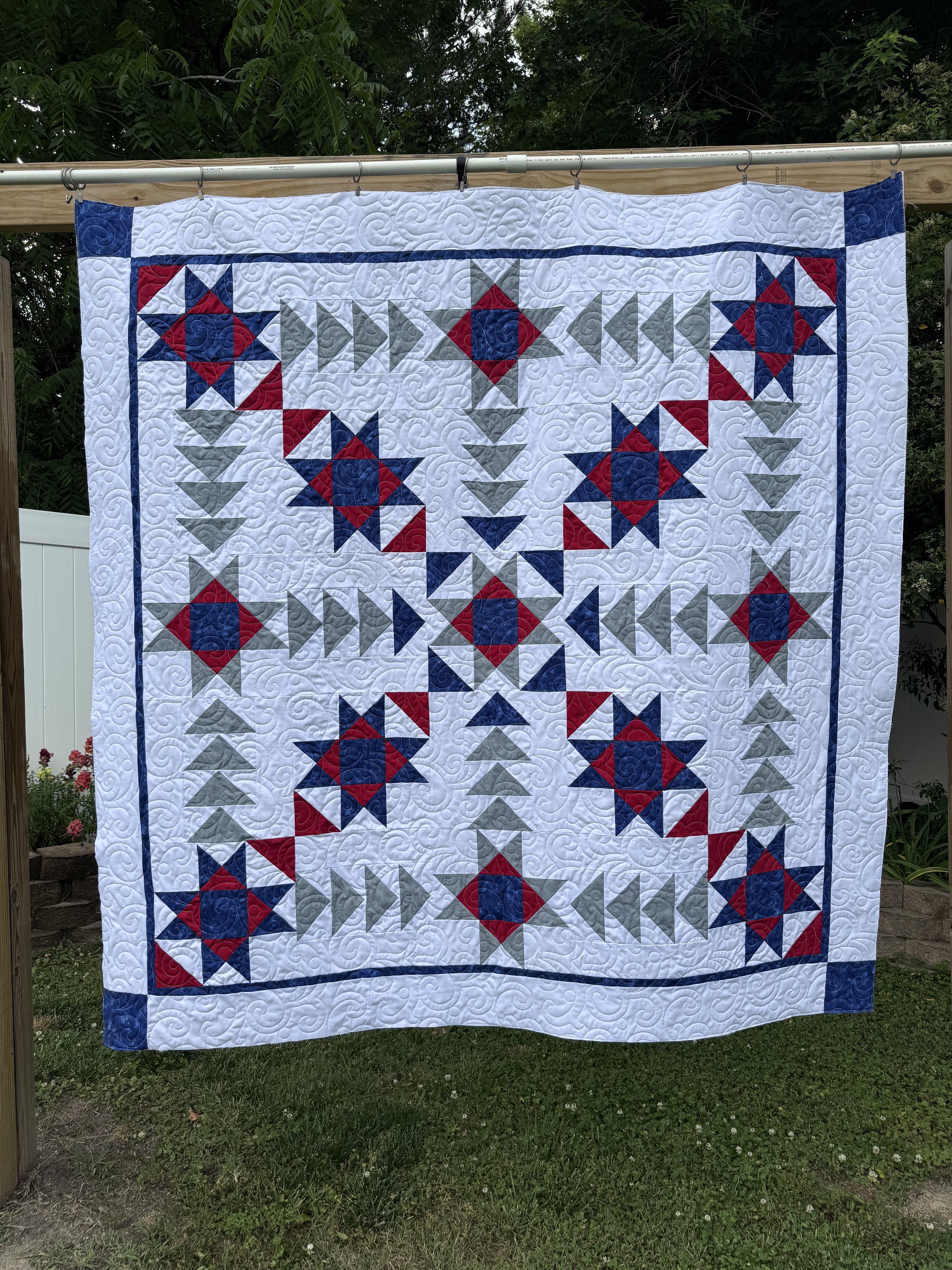 A quilt hanging on a clothesline in a backyard, featuring a patriotic red, white, and blue star pattern with a border.