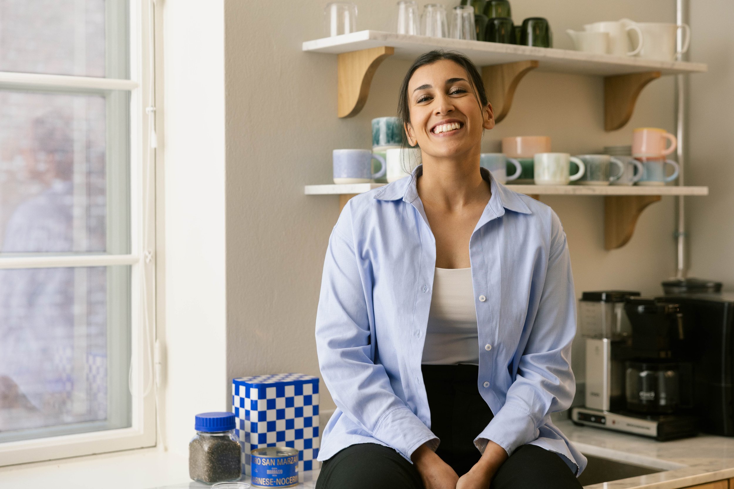 A woman with dark hair smiling in a kitchen, sitting near a window with a white frame. Brand photgraphy for banking.