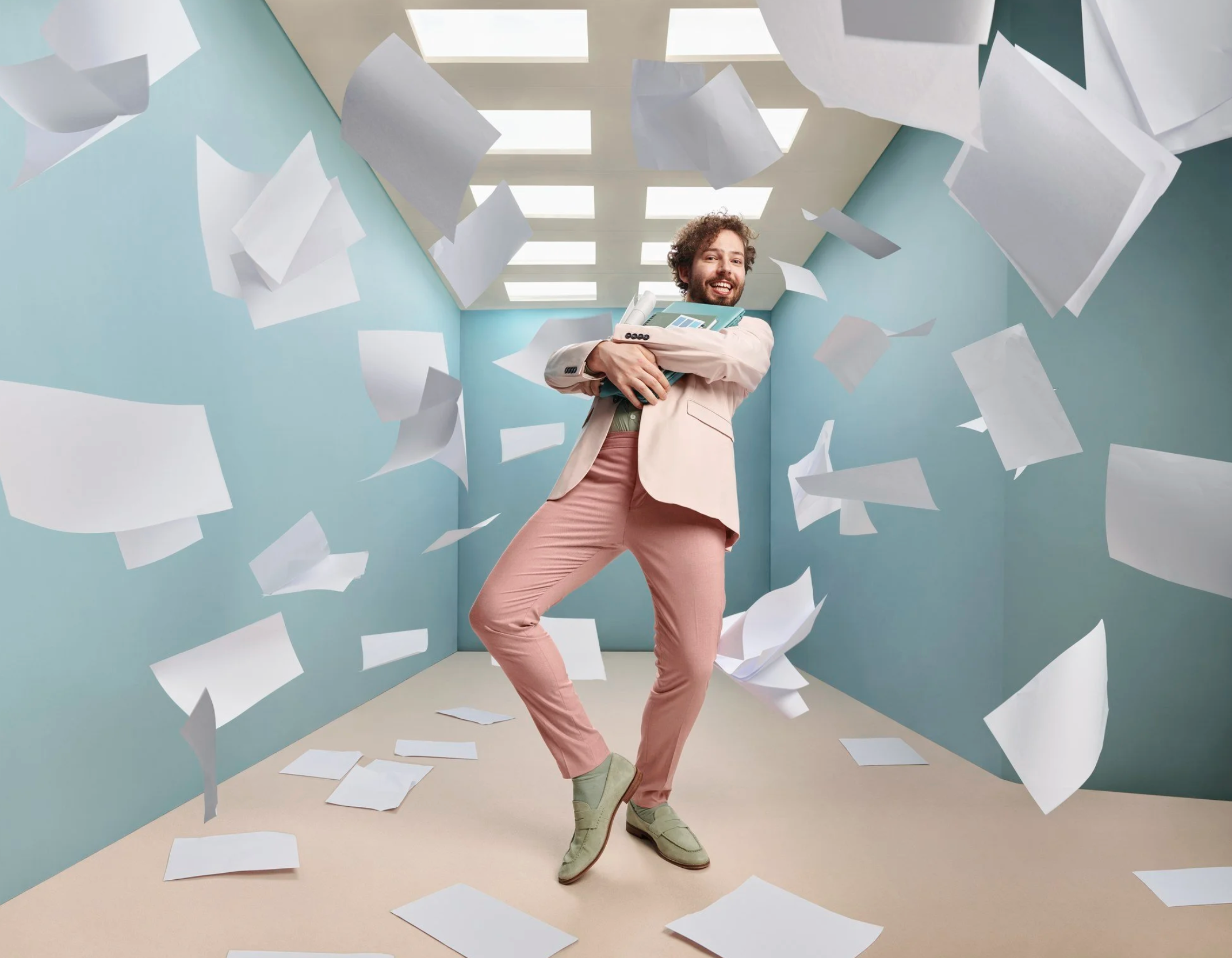 A man with curly hair and a beard, smiling and standing confidently while holding folders, surrounded by flying papers in a pastel-colored room with bright lighting. Advertising photography.