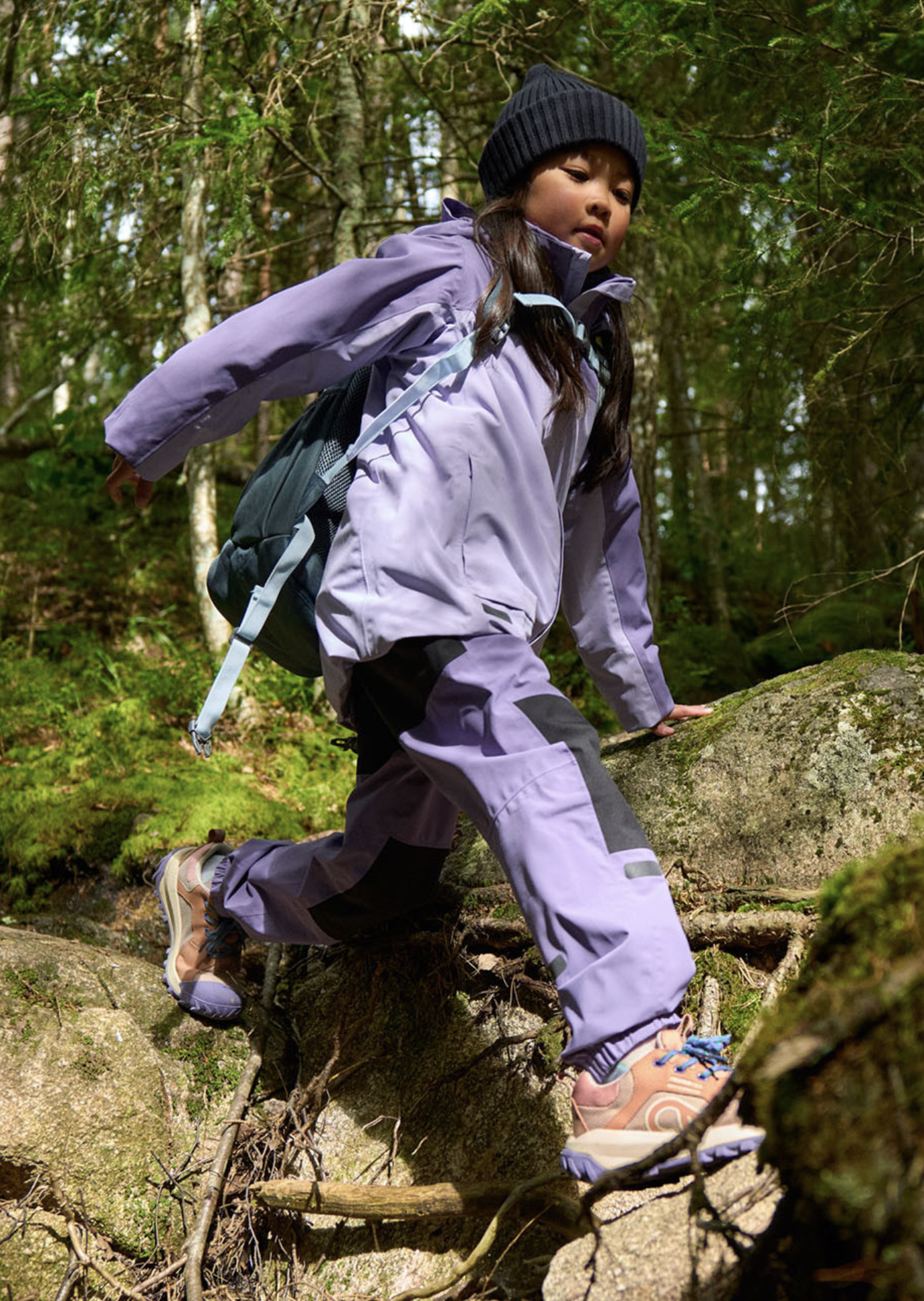A young girl climbing over rocks and fallen branches in a forest with green trees and sunlight filtering through the leaves. Ad photo for Reima.