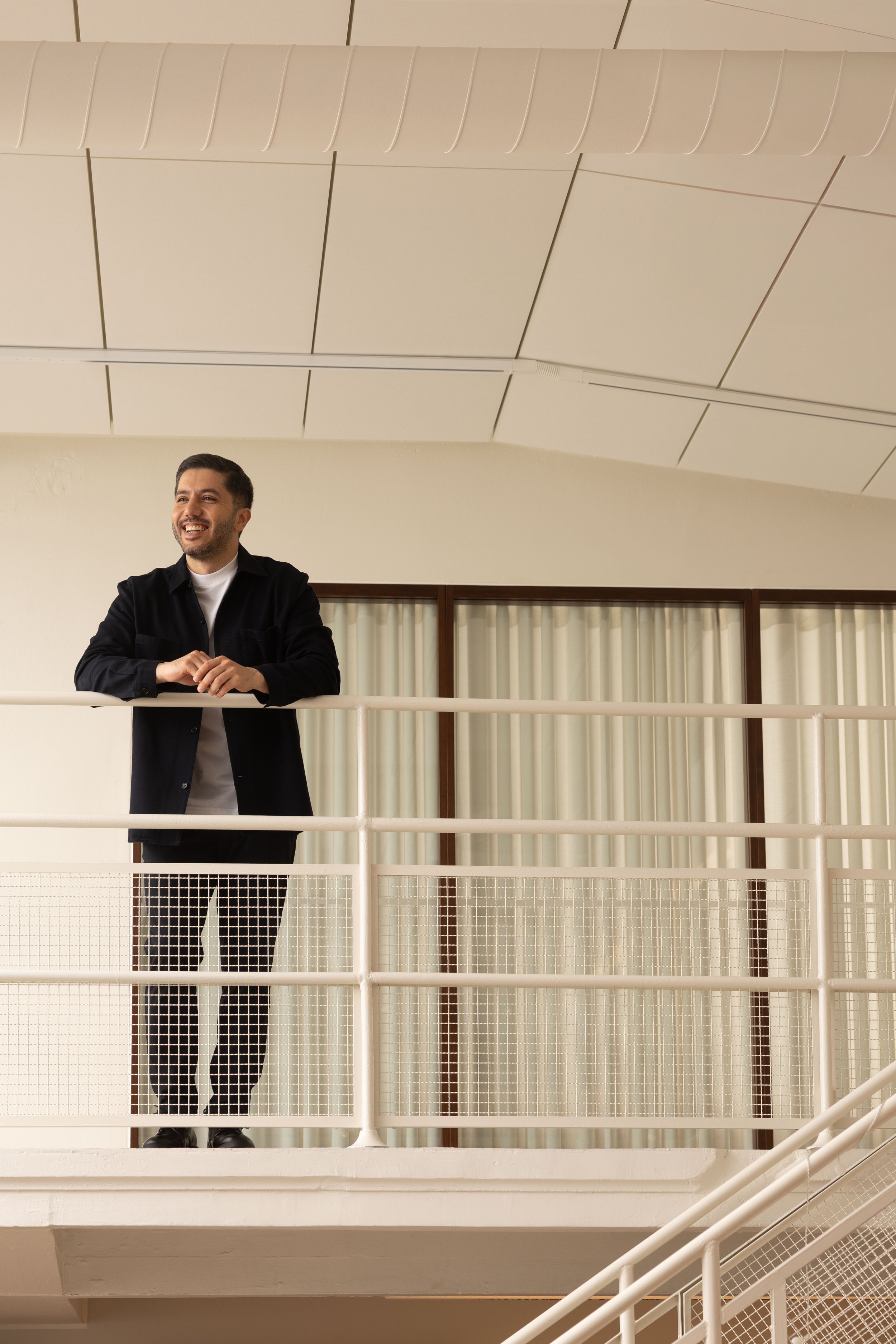 A man with dark hair, a beard, and a big smile stands with his arms crossed, leaning on a white railing inside a building. Brand photgraphy for banking.