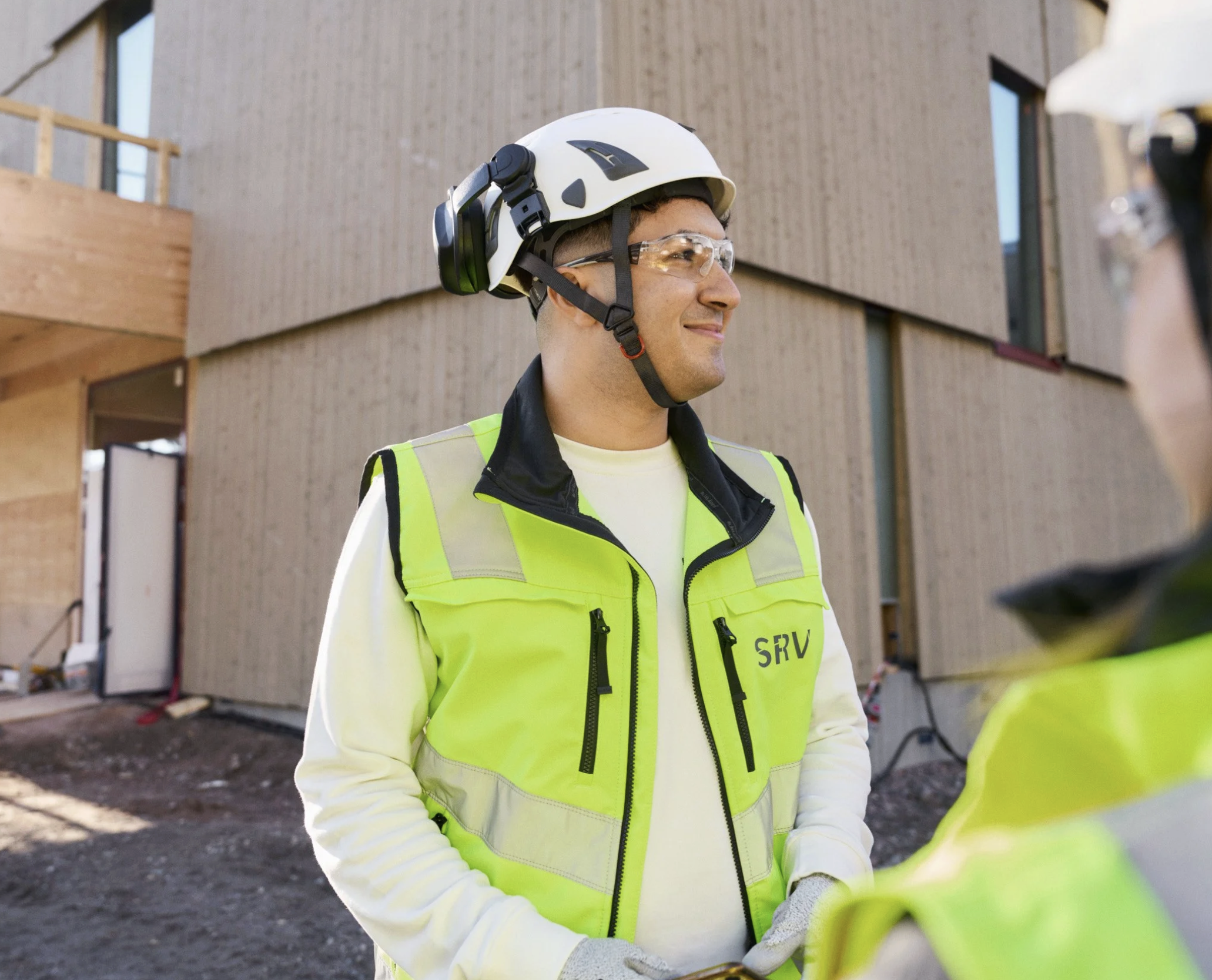 A construction worker in safety gear, including a white helmet, clear safety glasses, and a neon yellow safety vest, standing outdoors at a building site with a brown building in the background.