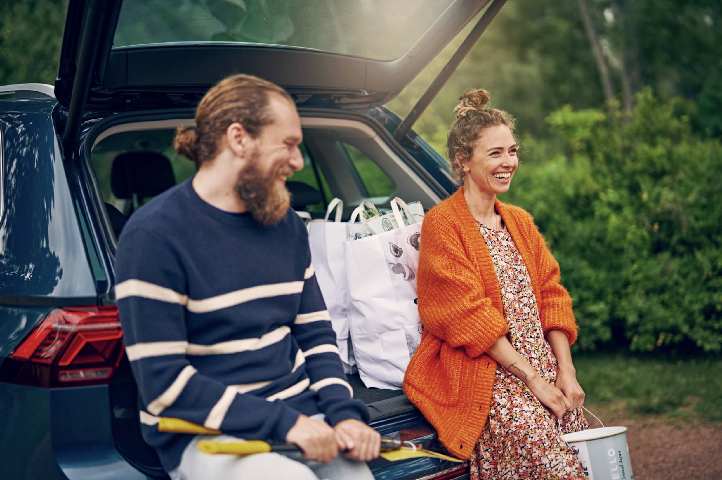A man and a woman sitting on the edge of a car trunk with groceries, smiling and enjoying an outdoor setting with greenery in the background. Brand photography for a hardware store chain.