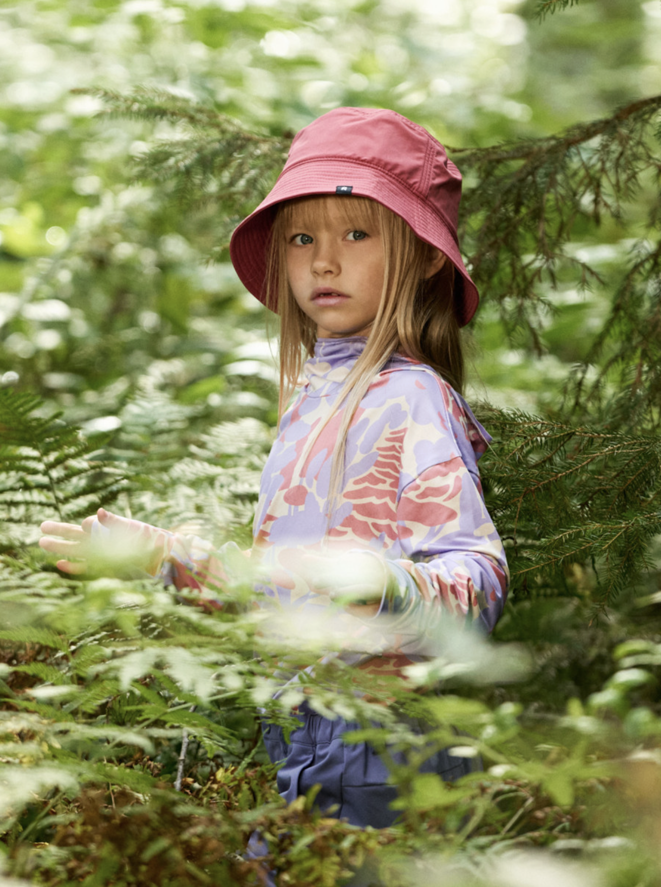 A young girl with long blonde hair, stands among green foliage in a forest. Ad photo for Reima.