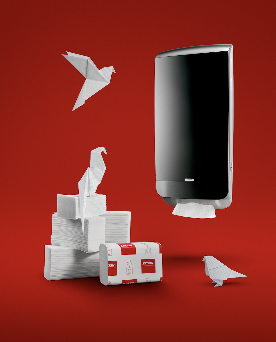A black paper towel dispenser mounted on a red background, with origami birds sitting on tissue stacks and flying nearby. Hybrid photo using traditional photgraphy and AI. Ad phography.