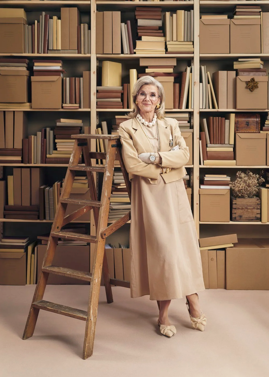 A senior woman in beige clothing stands in front of a large bookshelf filled with books and storage boxes, accompanied by a wooden ladder.  Advertising photography.