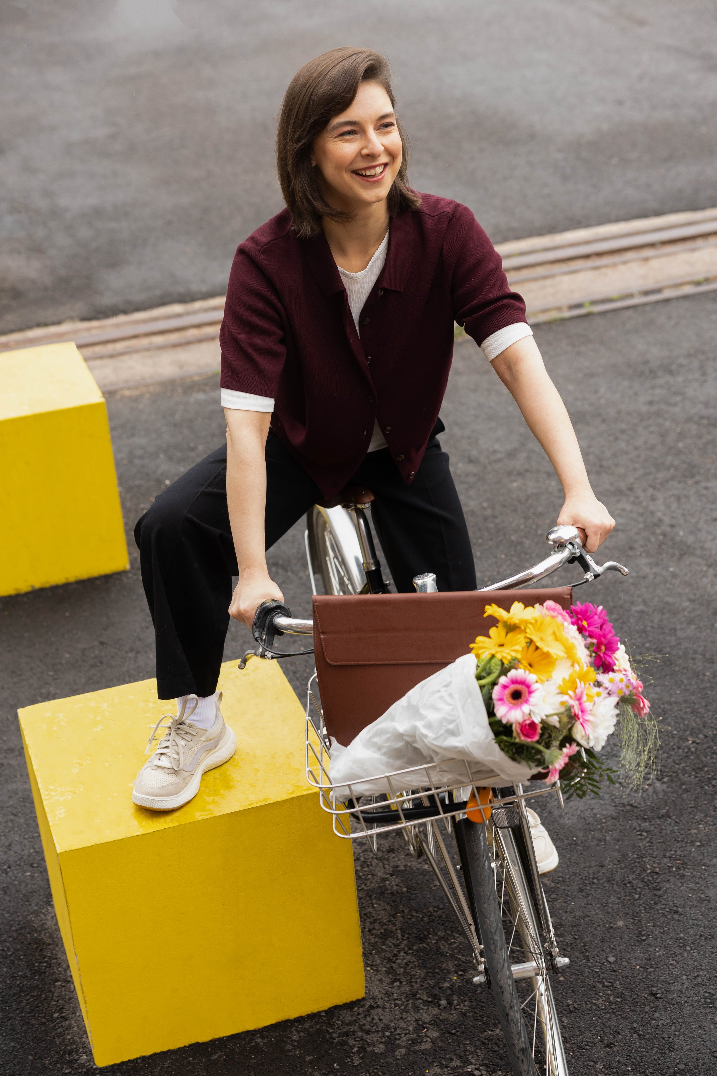A woman riding a bicycle with a basket of flowers, standing on a yellow block on a street. Brand photgraphy for banking.