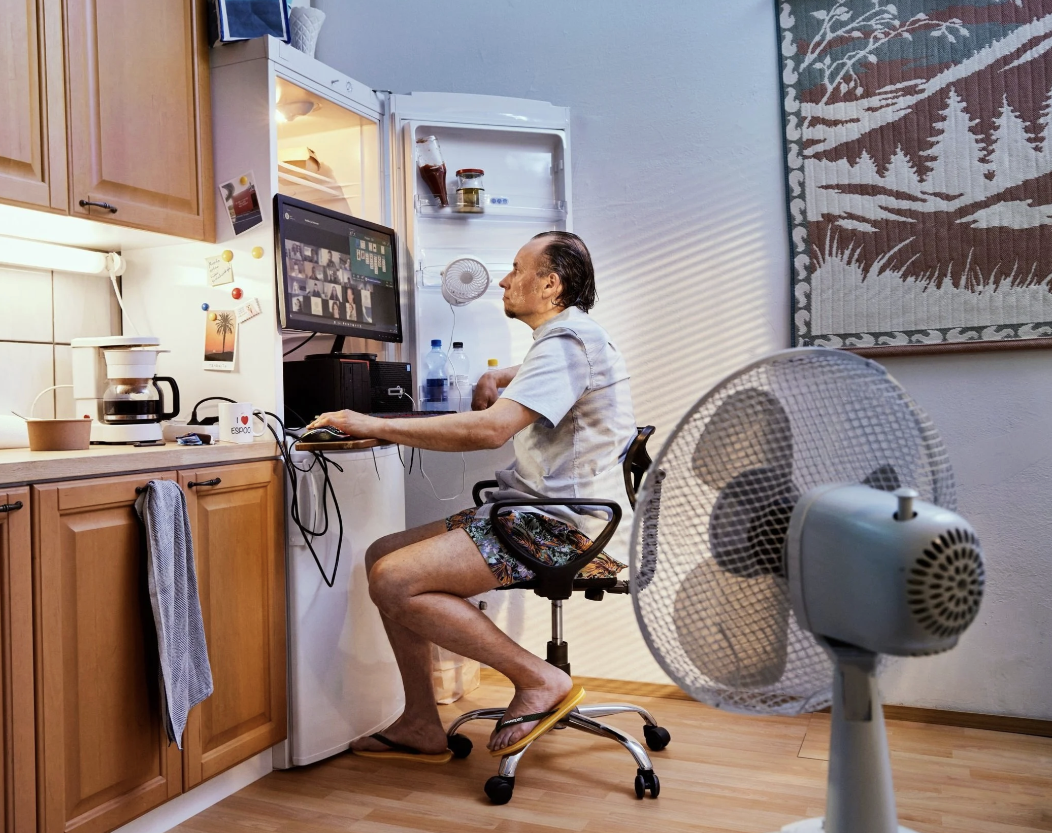 A man sitting in a kitchen working on a computer at a desk wearing flip-flops, shorts, and a t-shirt. Ad photography showcasing cooling systems.