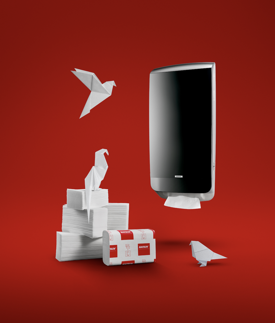 A black paper towel dispenser mounted on a red background, with origami birds sitting on tissue stacks and flying nearby. Hybrid photo using traditional photgraphy and AI.