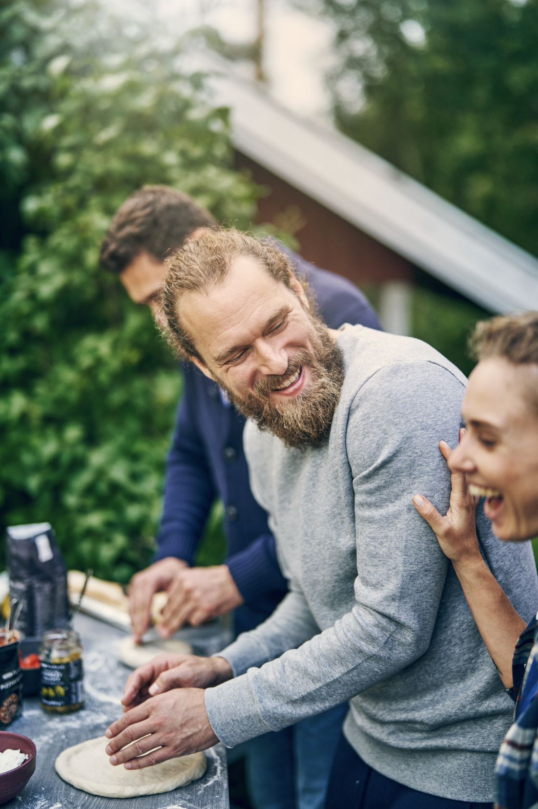 A group of people making pizza outdoors, smiling and laughing together.