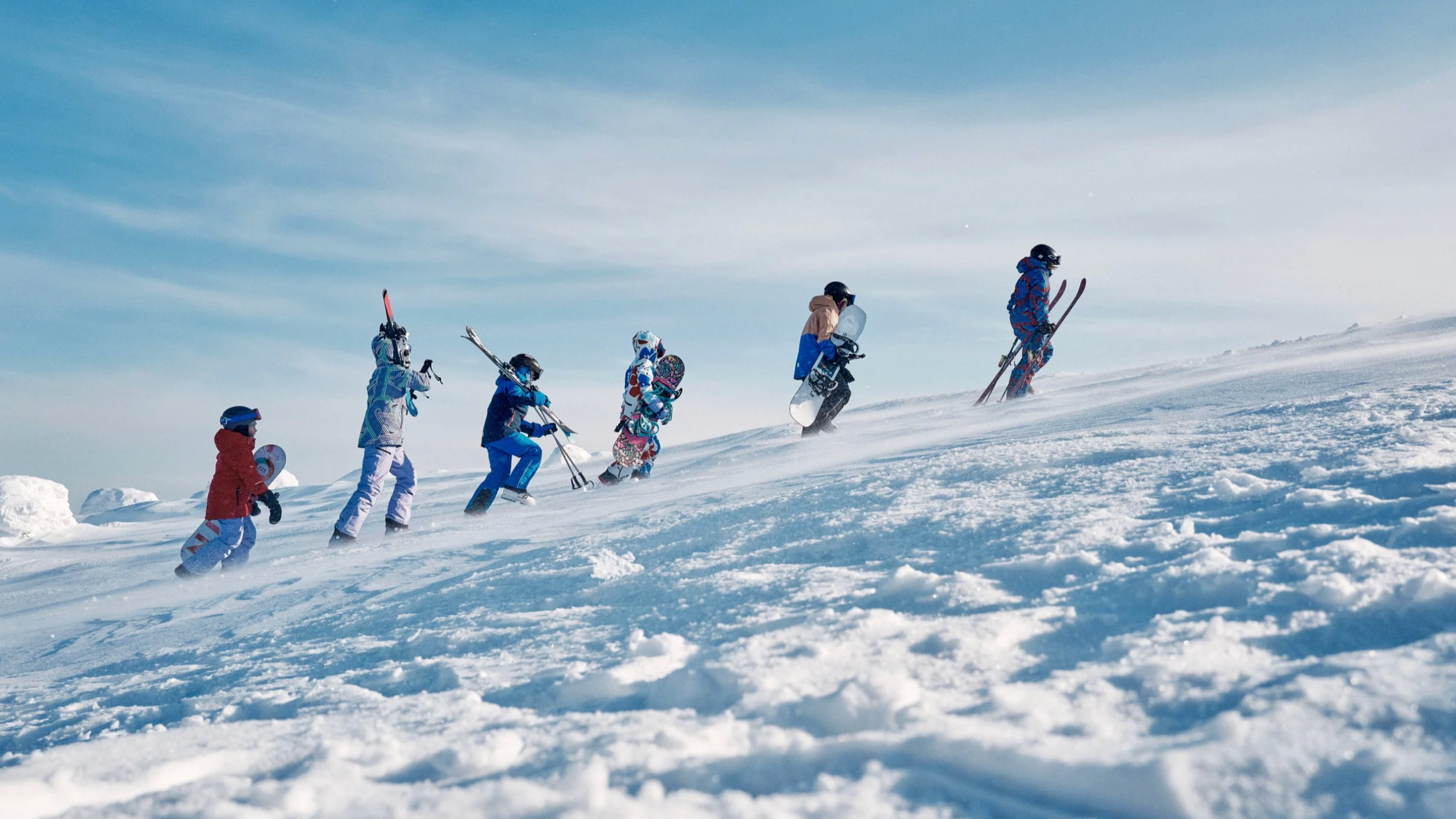 Children walking up a snowy slope with ski gear, under a blue sky. Ad photo for Reima.