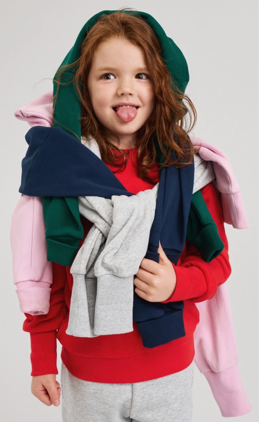 A young boy with red hair making a funny face with his tongue sticking out, wearing a red sweatshirt, and carrying multiple colorful hoodies layered on his shoulders. Ad photo for Reima.