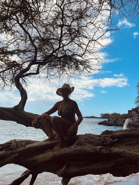Kaja mit Hut sitzt auf einem knorrigen Baum am Strand von Hawaii