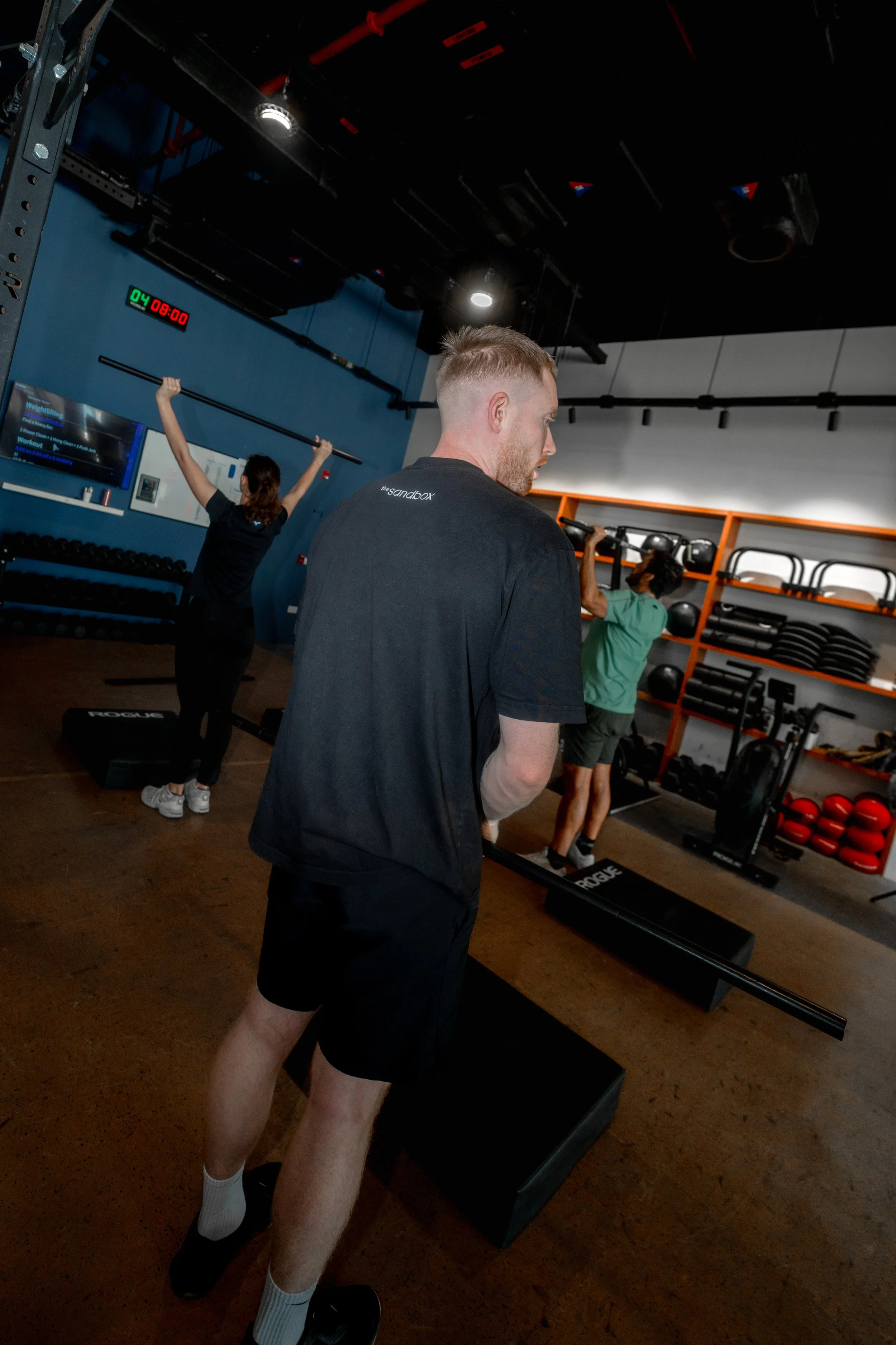 People working out in a gym, with one person lifting a barbell in the background and another person in front, wearing black shorts, a black t-shirt, white socks, and black shoes.