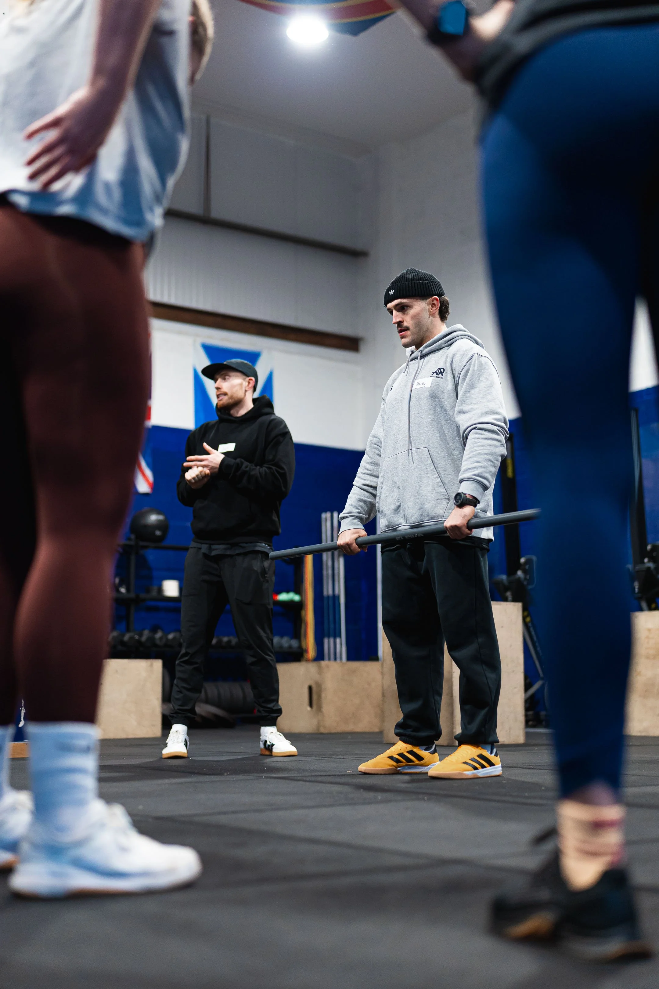 People in a gym listening to a trainer, focus on a man wearing a gray hoodie, black beanie, and yellow sneakers.